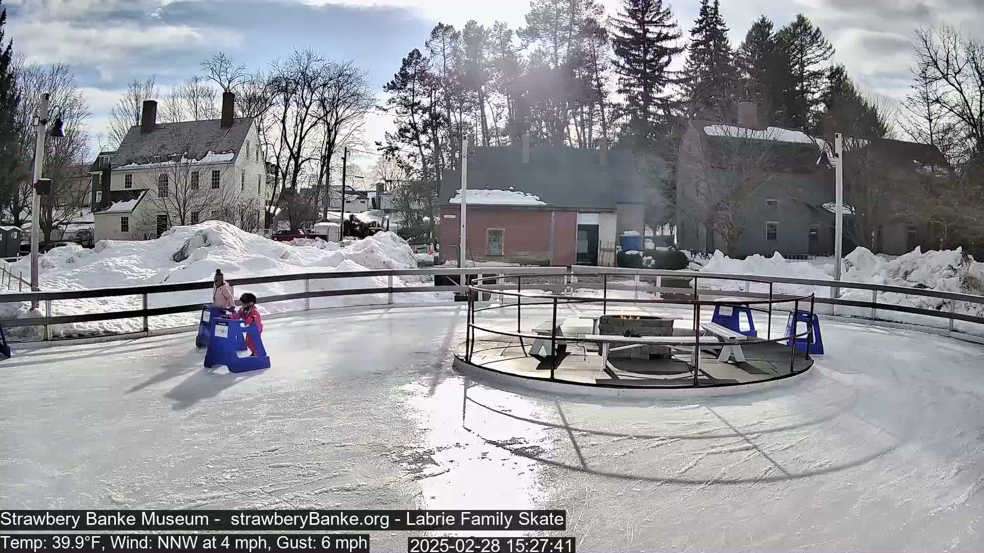 Two children on a snowy outdoor ice rink use plastic skating aids near a fire pit, under a mostly sunny sky.