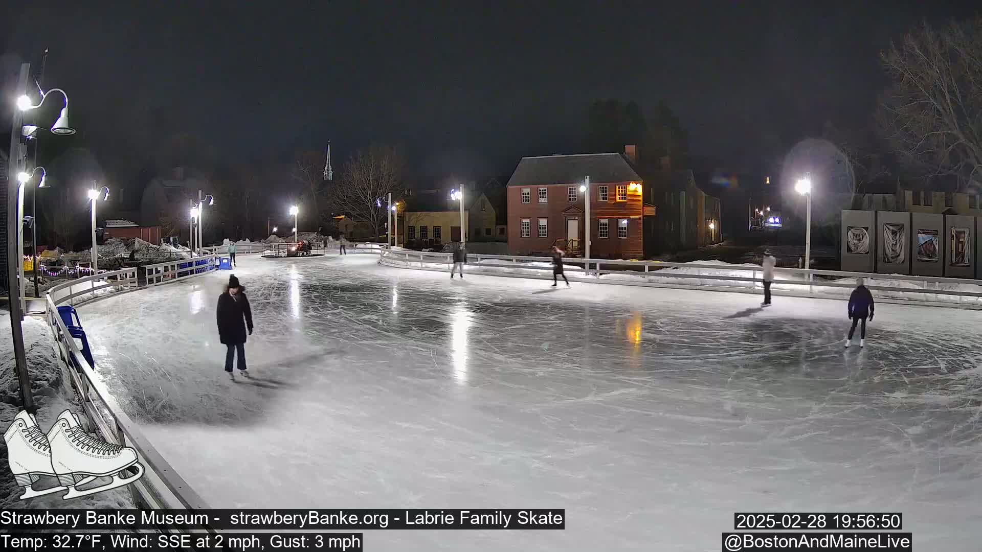 At night, several people ice skate on an outdoor rink illuminated by lights, under a dark sky with a slight wind.