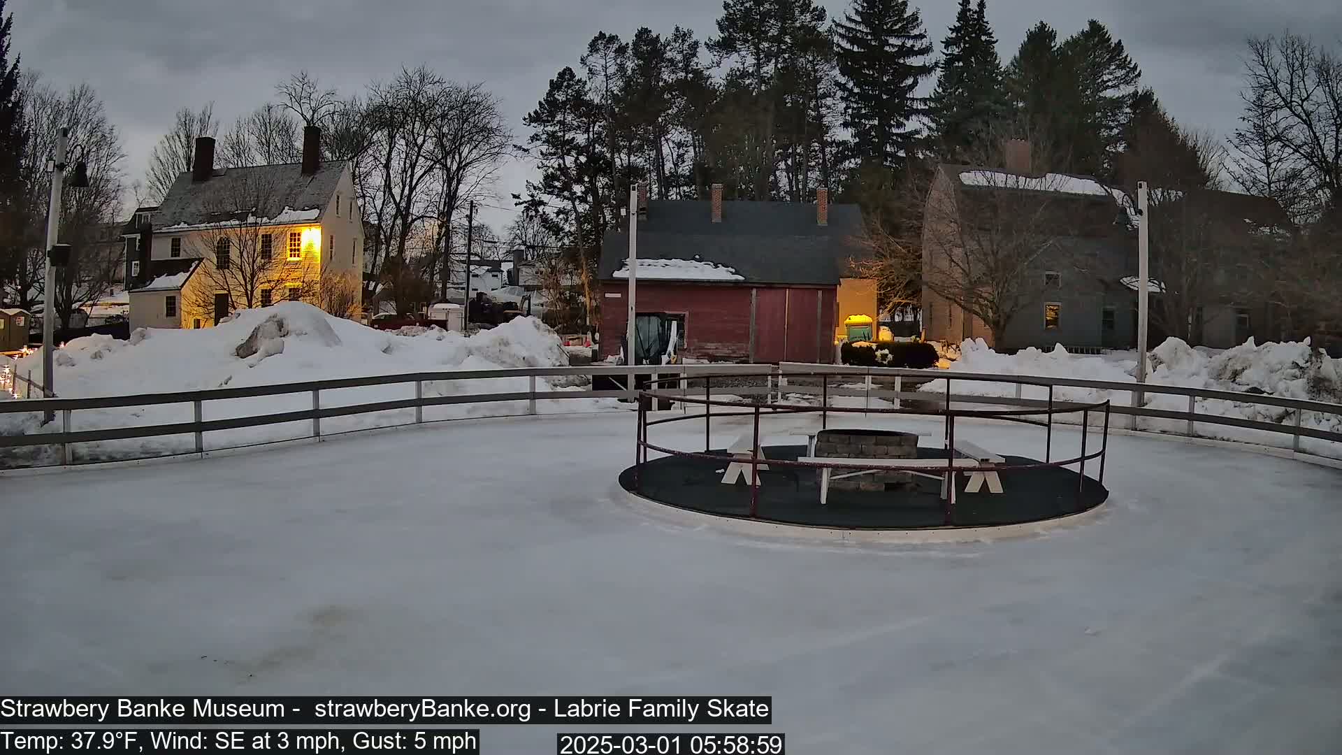A snow-covered outdoor skating rink with a fire pit in the center, surrounded by historic-looking buildings under a twilight sky.