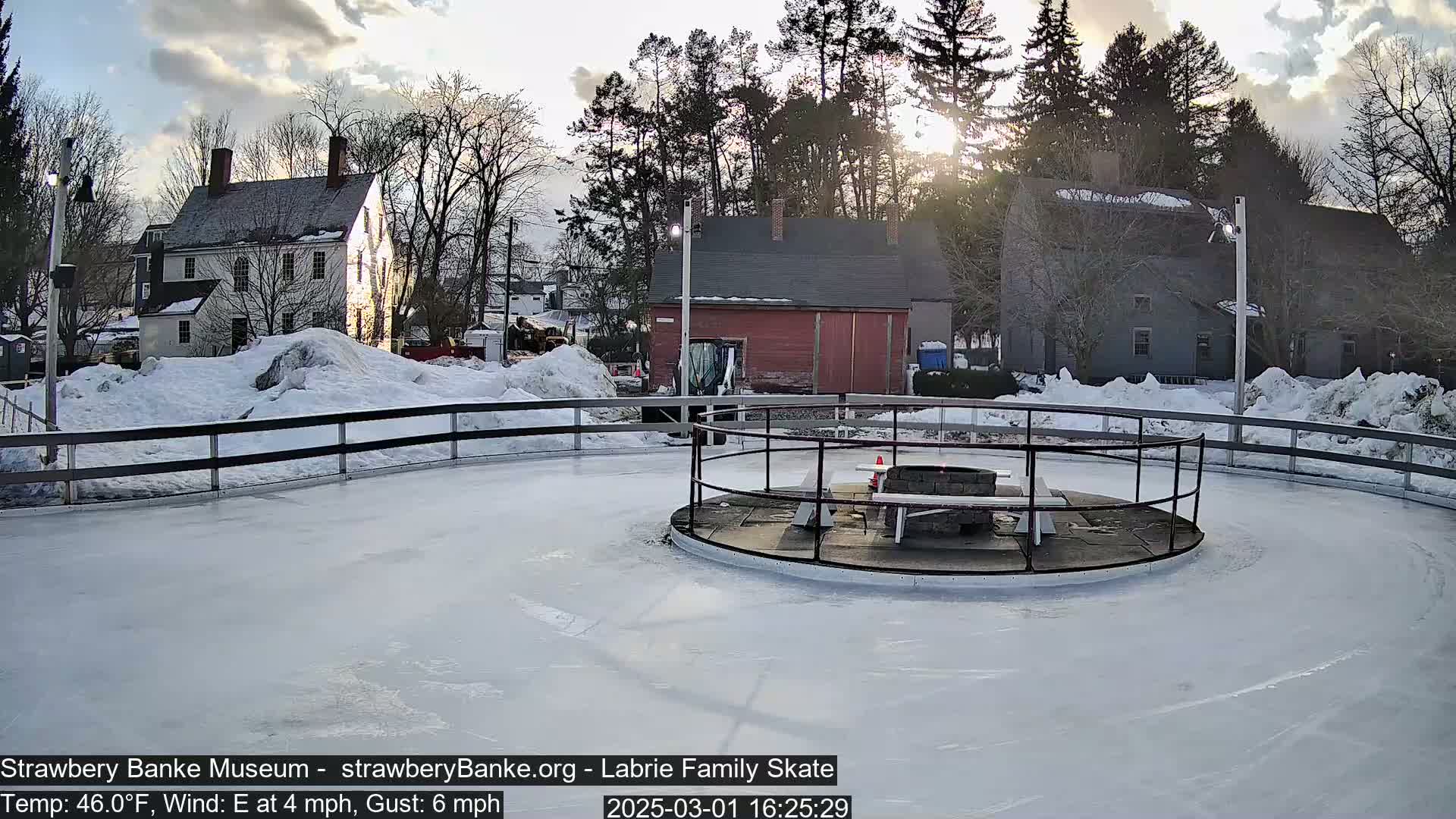 A partially sunny, cold day with snow on the ground surrounding a circular outdoor ice skating rink with a central fire pit and benches.