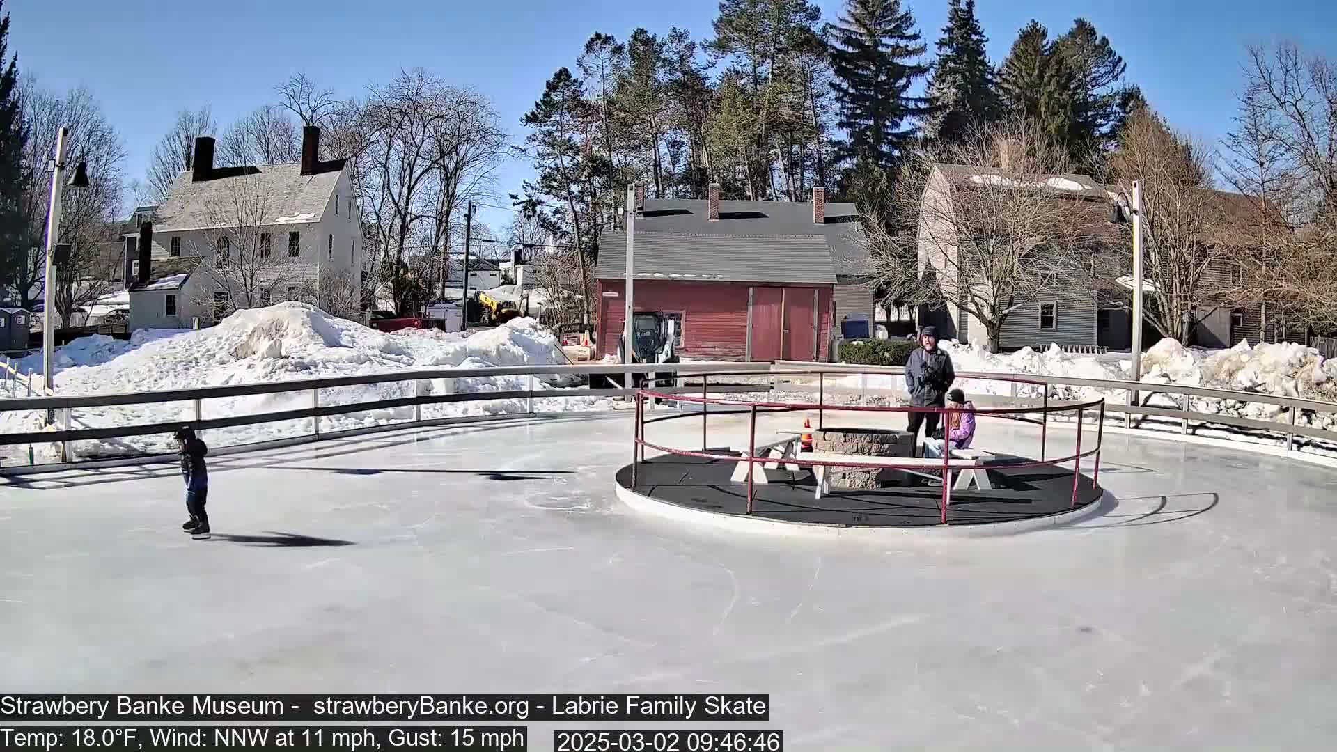 A sunny, cold day shows a circular ice skating rink with a few skaters and onlookers near a fire pit, surrounded by snow-covered ground and historic-looking buildings.