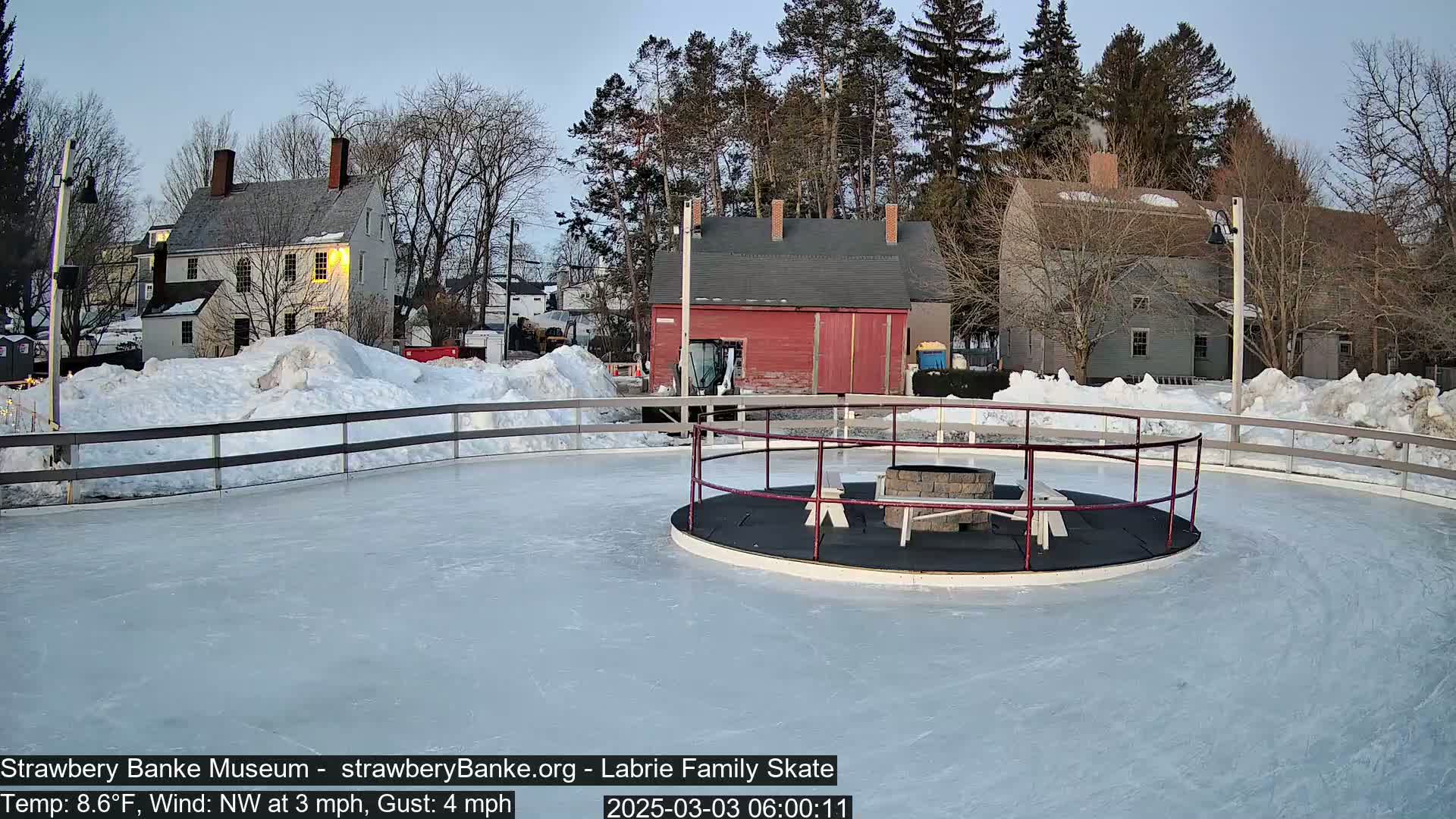 A frozen outdoor skating rink with a fire pit in the center, surrounded by snow and historic-looking buildings under a twilight sky.