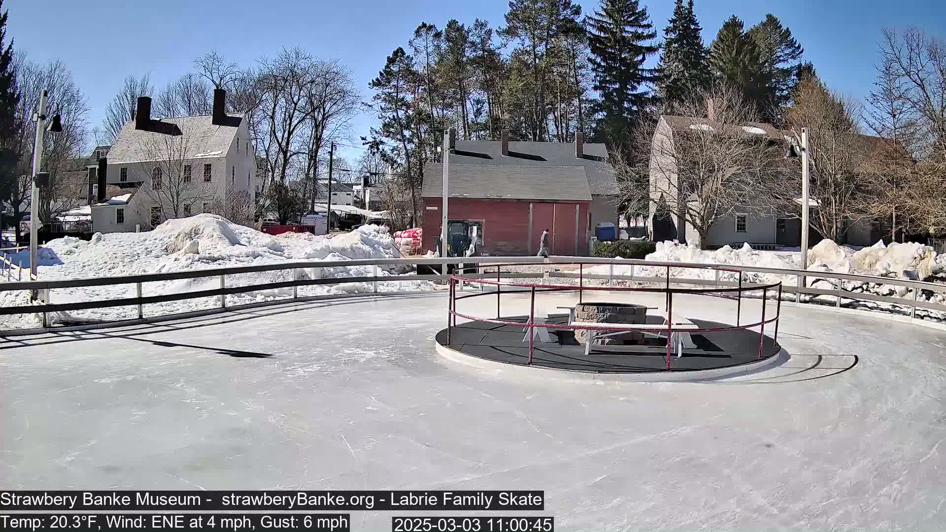 A sunny outdoor ice rink with a fire pit in the center, surrounded by snow and historic-looking buildings.