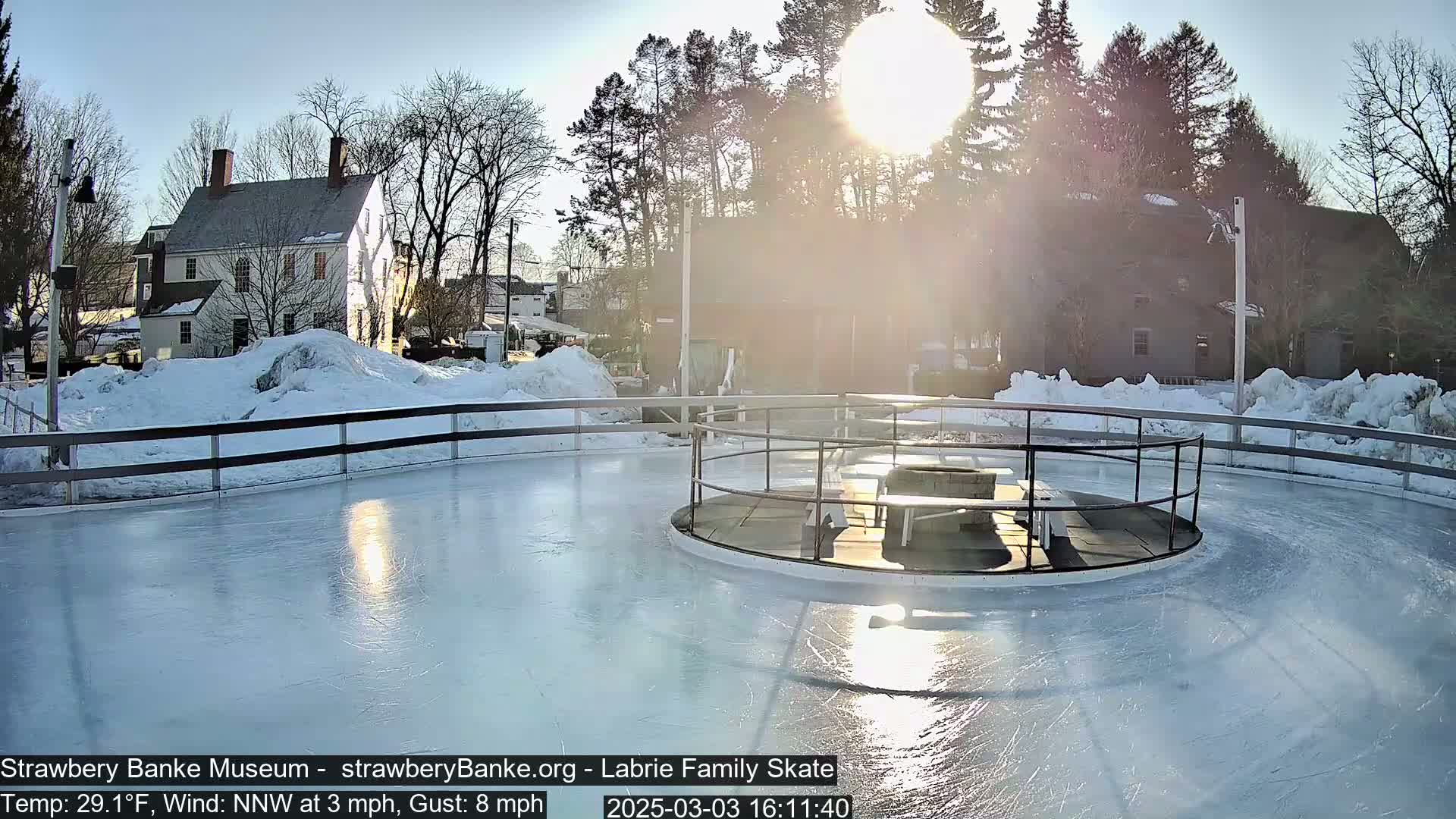 A sunny, slightly windy day shows an outdoor ice skating rink partially surrounded by snow, with a building visible in the background.