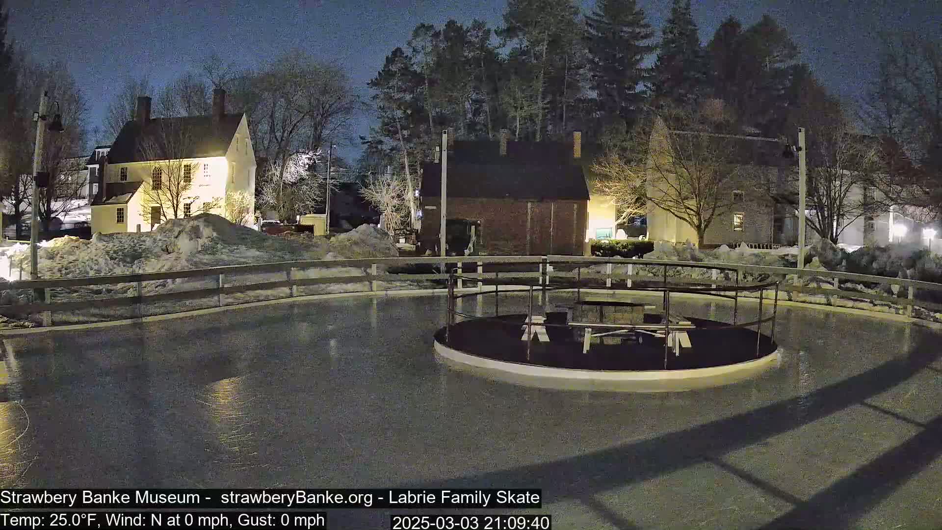 A nighttime view of an outdoor ice rink surrounded by snow-covered ground and historical-looking buildings under a clear, dark sky.