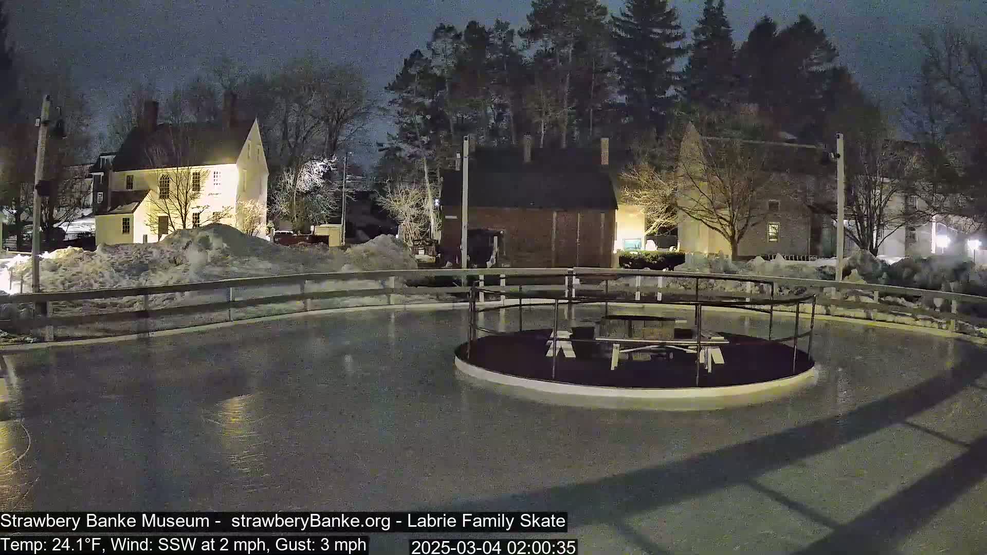 An outdoor ice rink at night, illuminated by artificial light, surrounded by snow-covered buildings and trees under a dark sky with a temperature of 24.1°F and a light south-southwesterly wind.