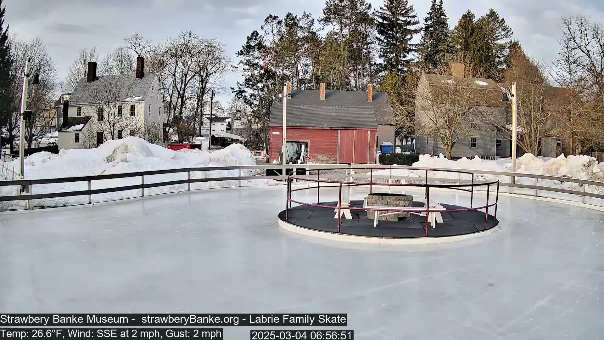 A snow-covered outdoor ice rink, surrounded by historical-looking buildings and trees, under a mostly cloudy sky.