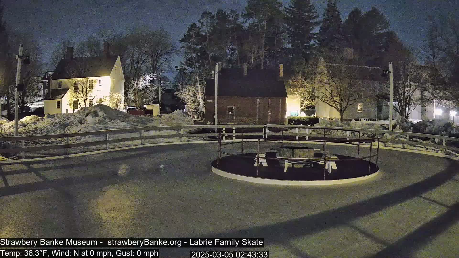 A nighttime view of a snow-covered outdoor skating rink surrounded by historic-looking buildings under a dark sky.