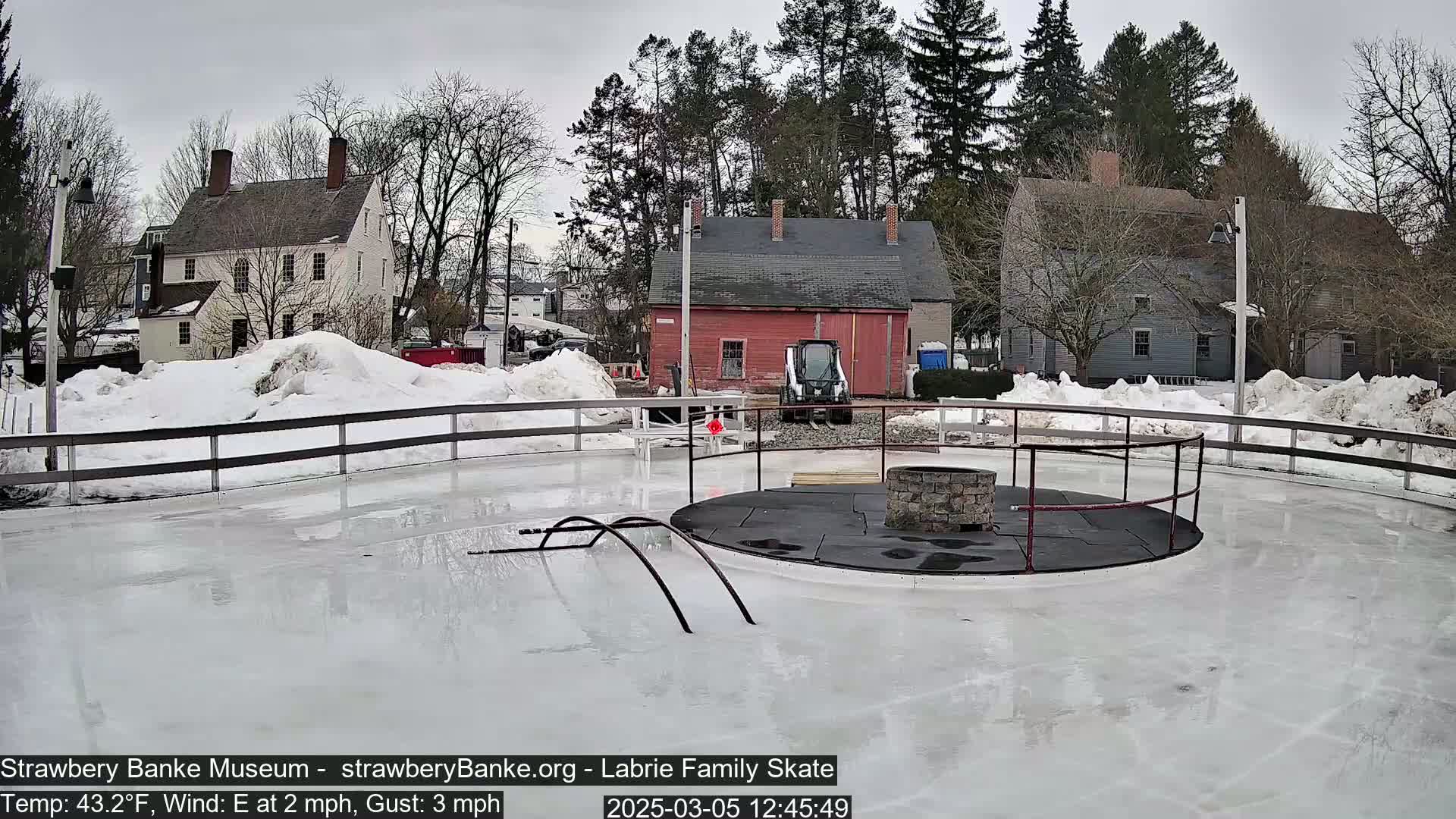 An outdoor ice skating rink, partially surrounded by snowdrifts, is situated near several historic-looking houses under an overcast sky with a light east wind.