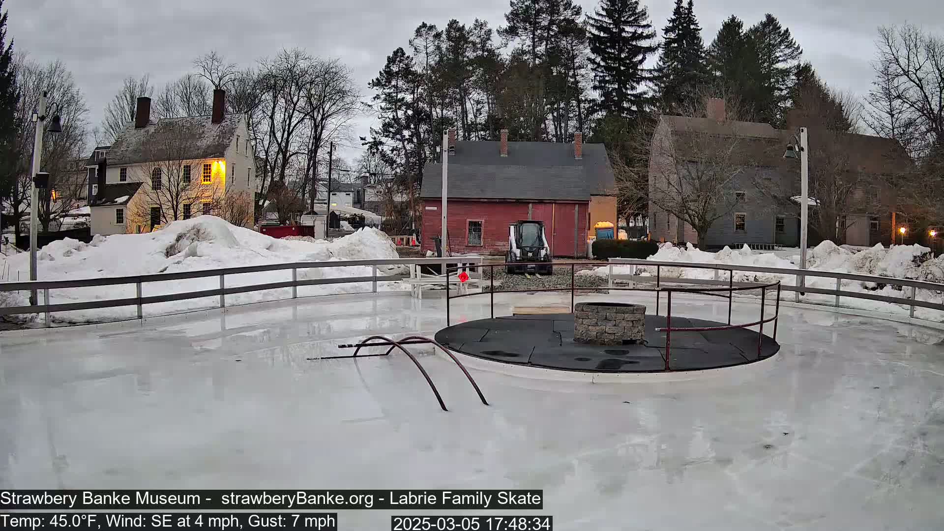 An outdoor ice rink with a fire pit in the center is surrounded by snow-covered ground and historic buildings under a cloudy sky.