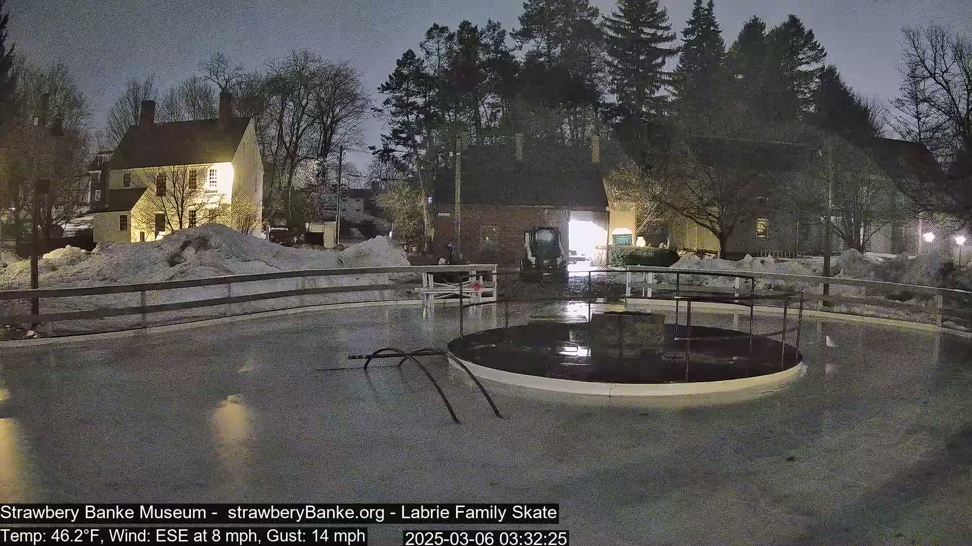 A nighttime view of a partially frozen skating rink next to a snow-covered colonial-style building under a dark sky with a wind from the east-southeast at 8 mph and gusts up to 14 mph.