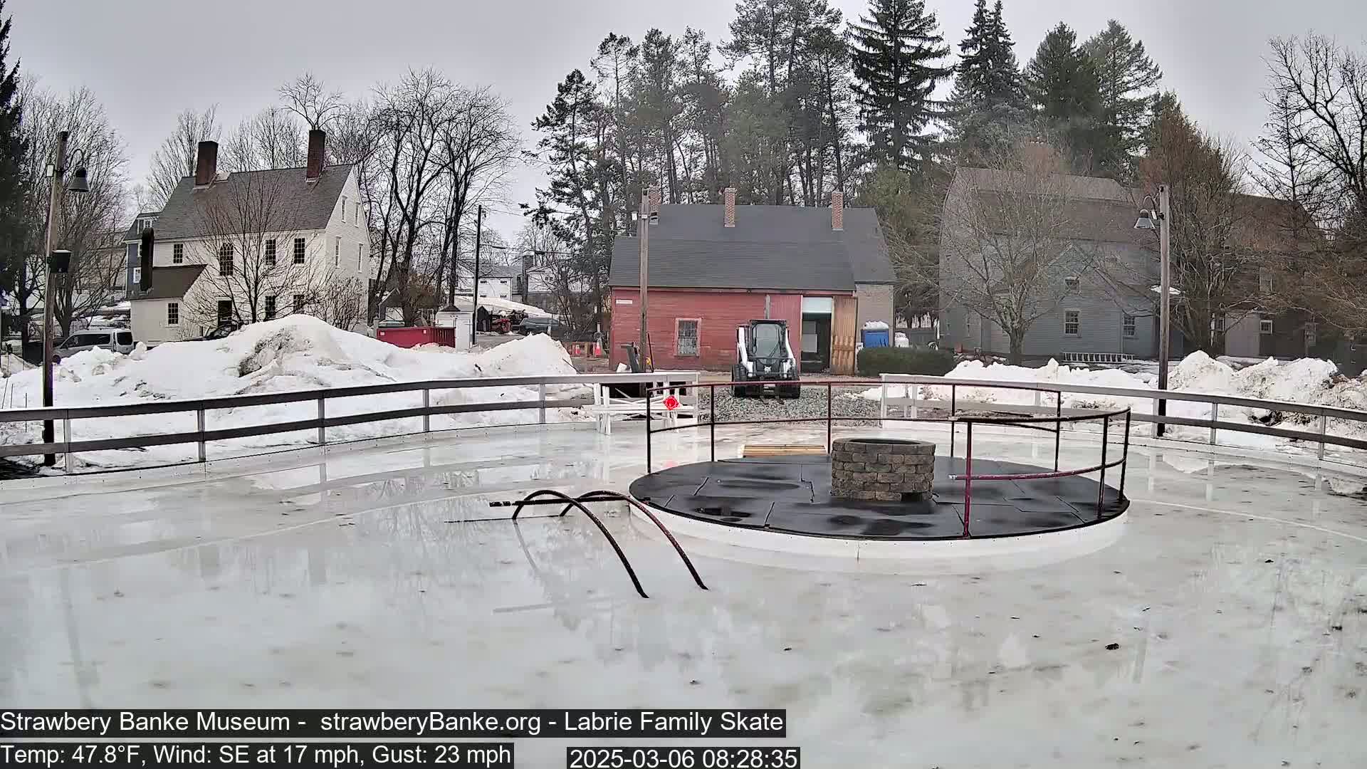 A partially melted ice rink with a central fire pit, surrounded by a low fence and snow-covered ground, on an overcast day with a southeasterly wind.