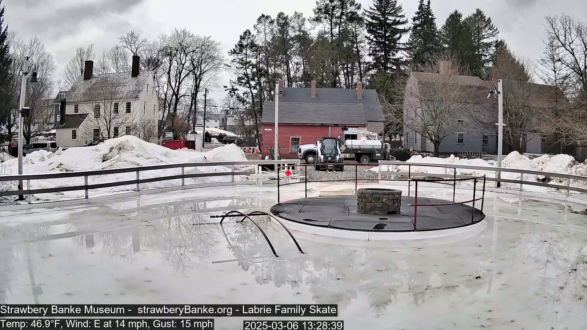 A mostly melted outdoor ice rink with a central fire pit, surrounded by snow and historic buildings under an overcast sky.