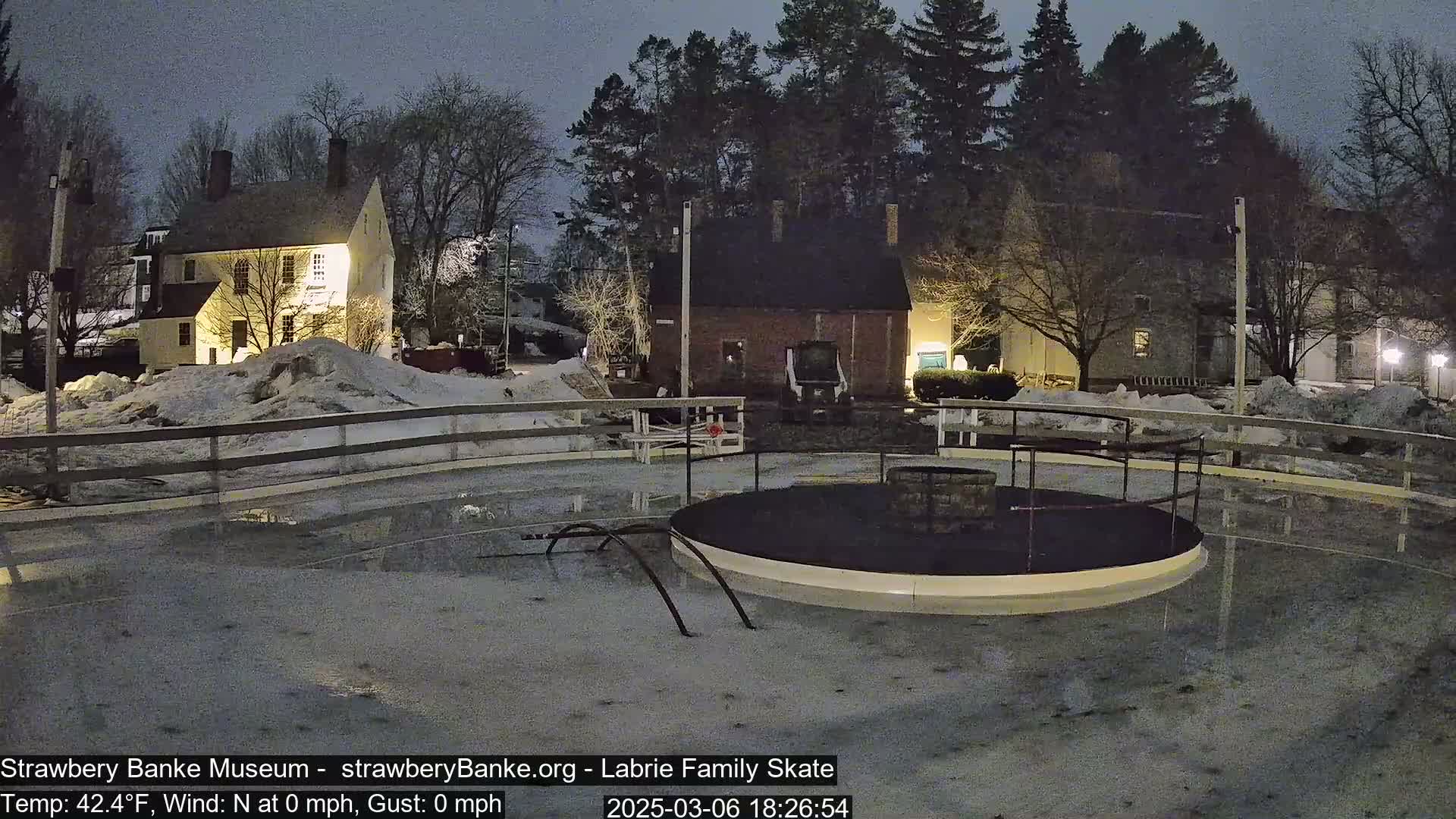 A nighttime view of a partially flooded ice rink, surrounded by snow-covered ground and illuminated buildings.