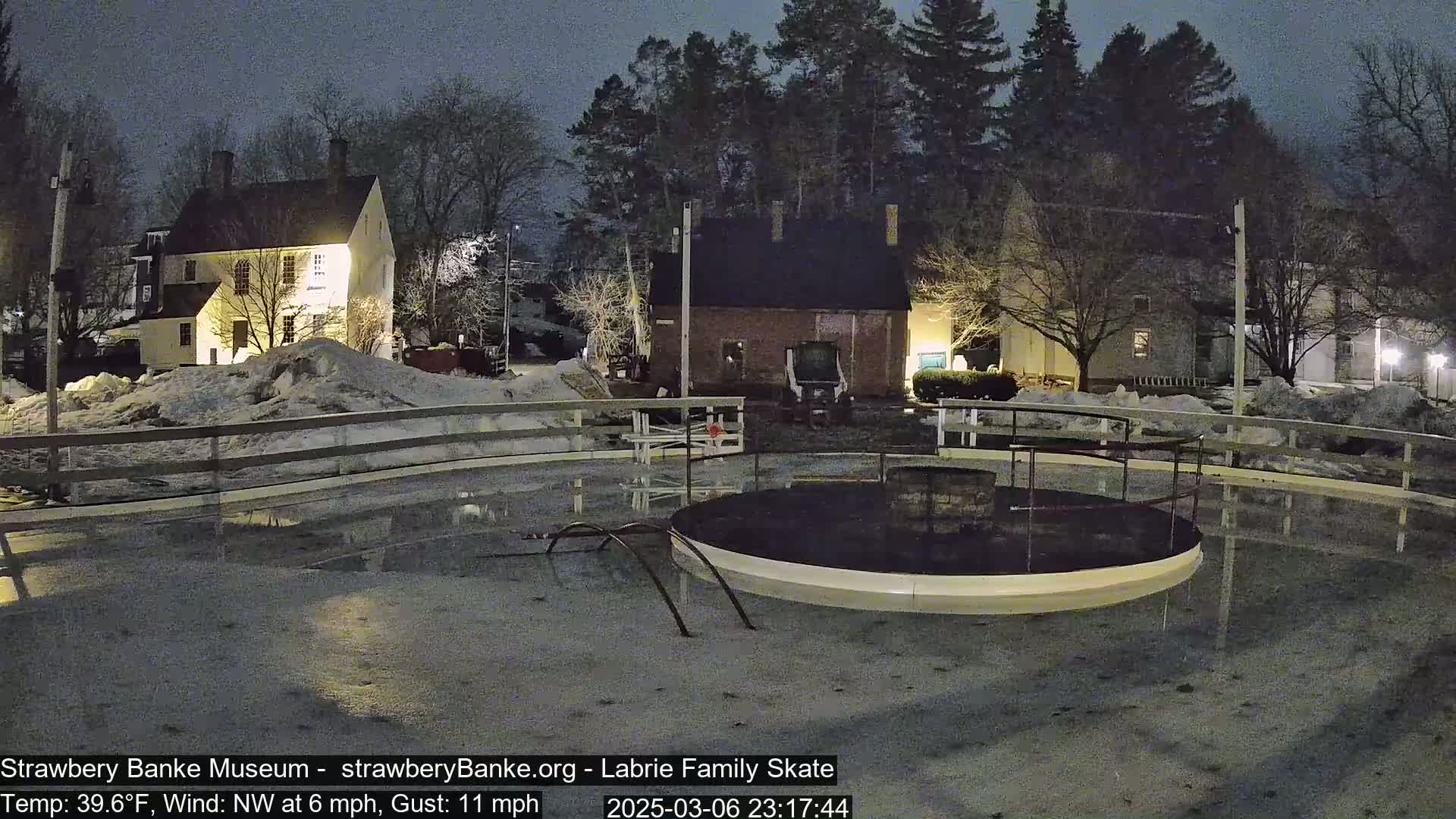 A nighttime view of a flooded ice rink surrounded by snow-covered ground and illuminated buildings under a dark sky.