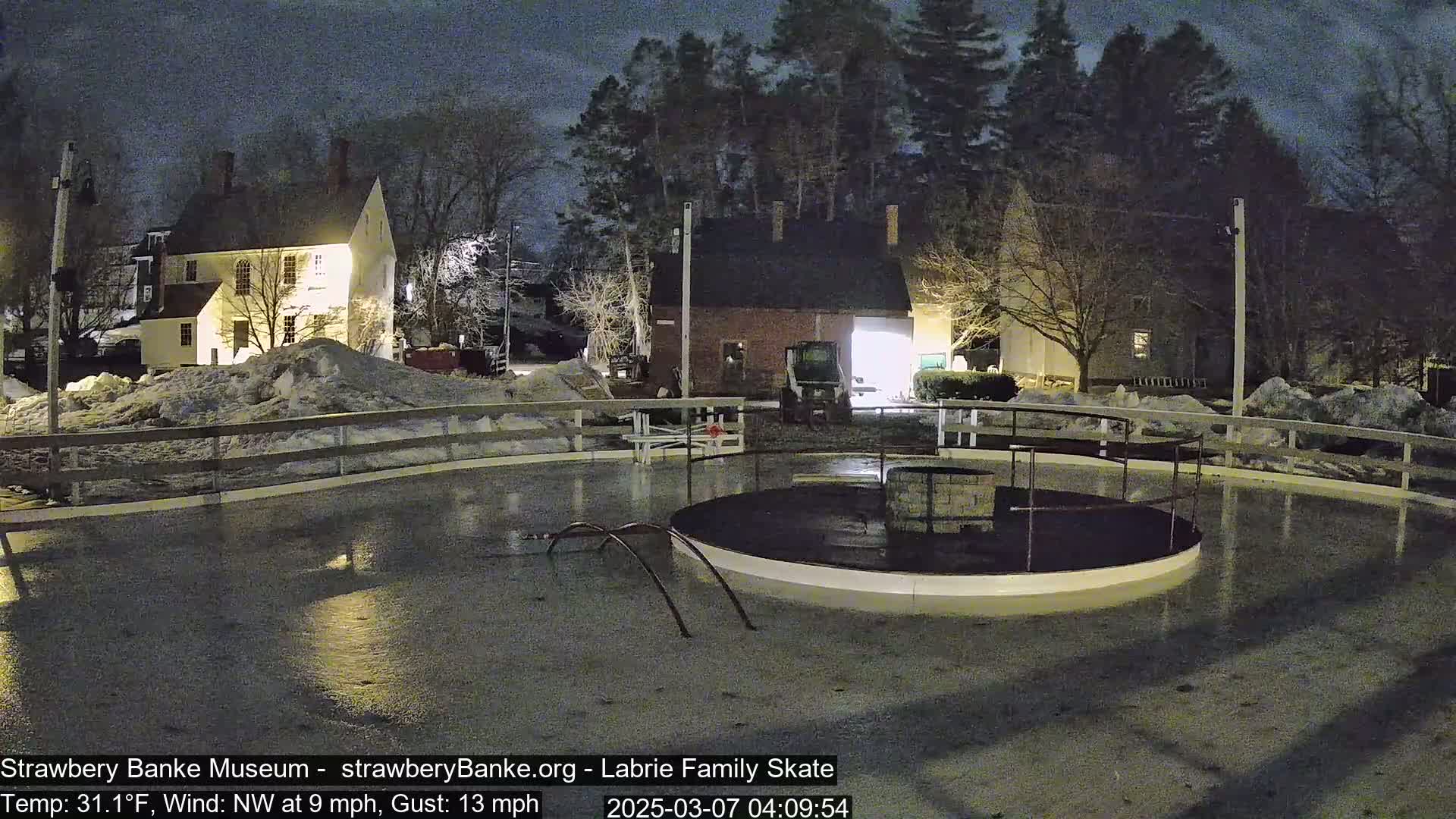 A wet, nighttime scene shows an ice skating rink surrounded by snow-covered ground and illuminated buildings under a dark sky.