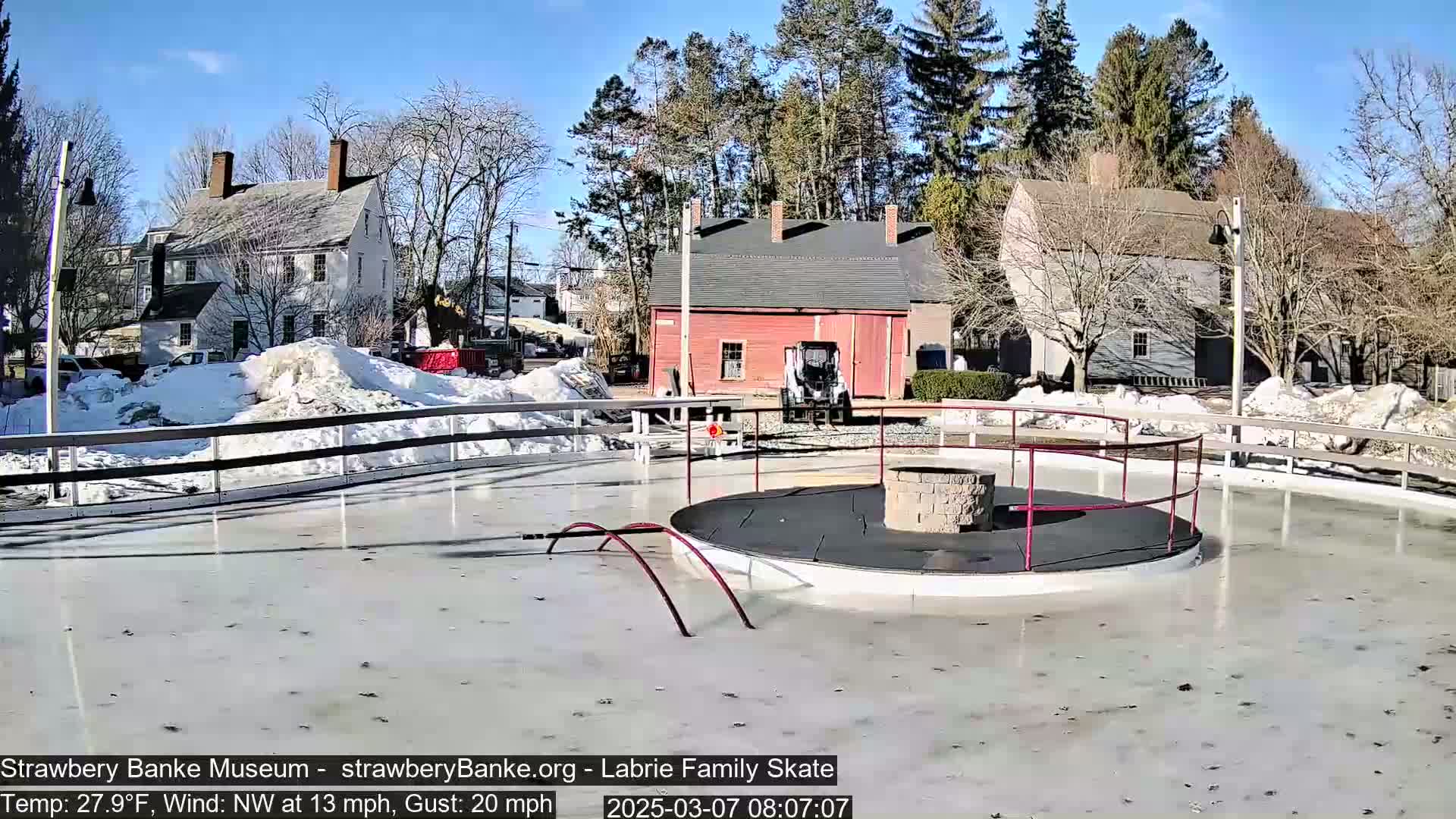 A partially melted ice rink with a central fire pit is surrounded by a low fence and historical-looking buildings under a sunny winter sky.