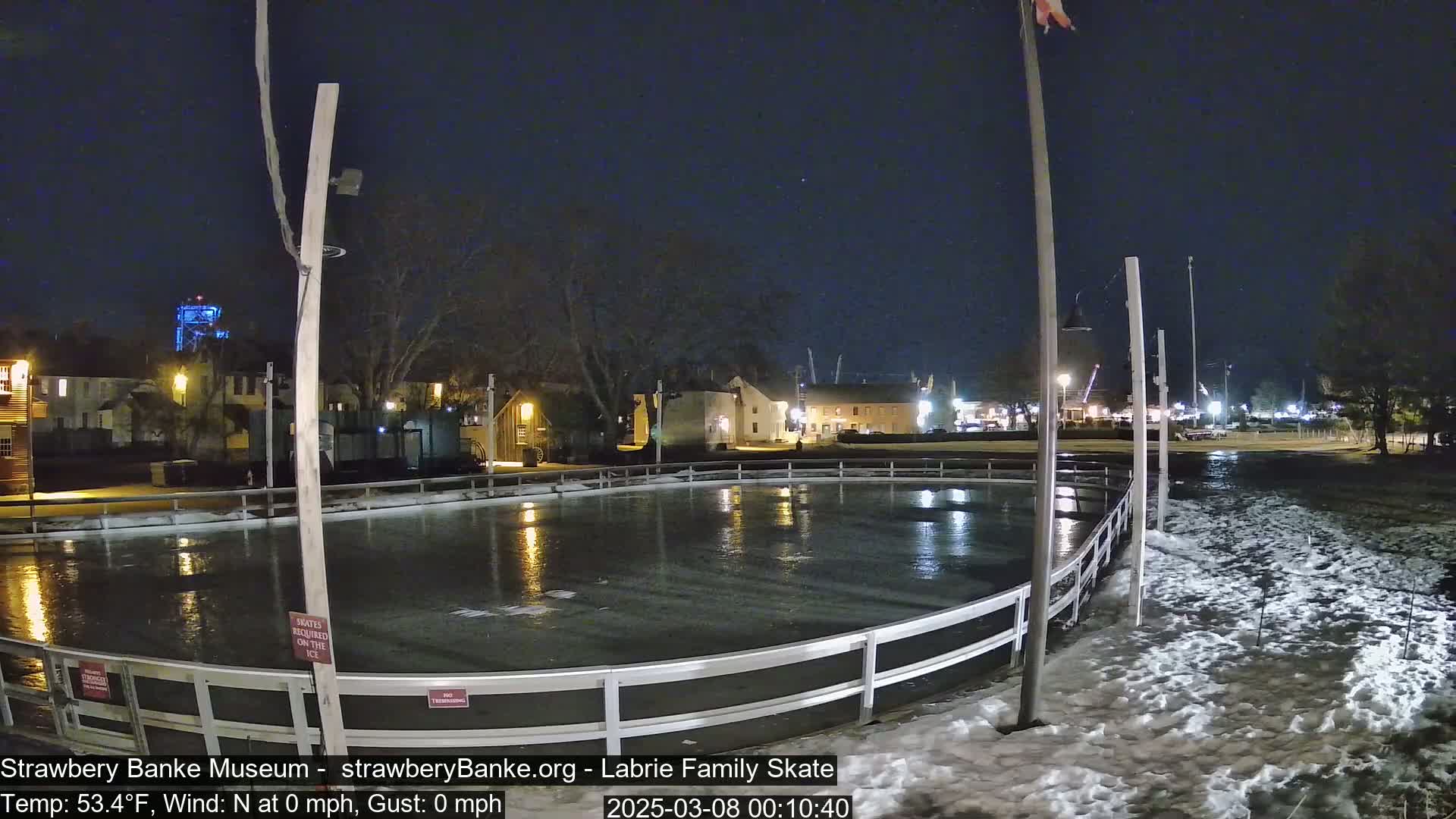An outdoor ice rink at night, surrounded by snow, with calm weather conditions and buildings visible in the background.