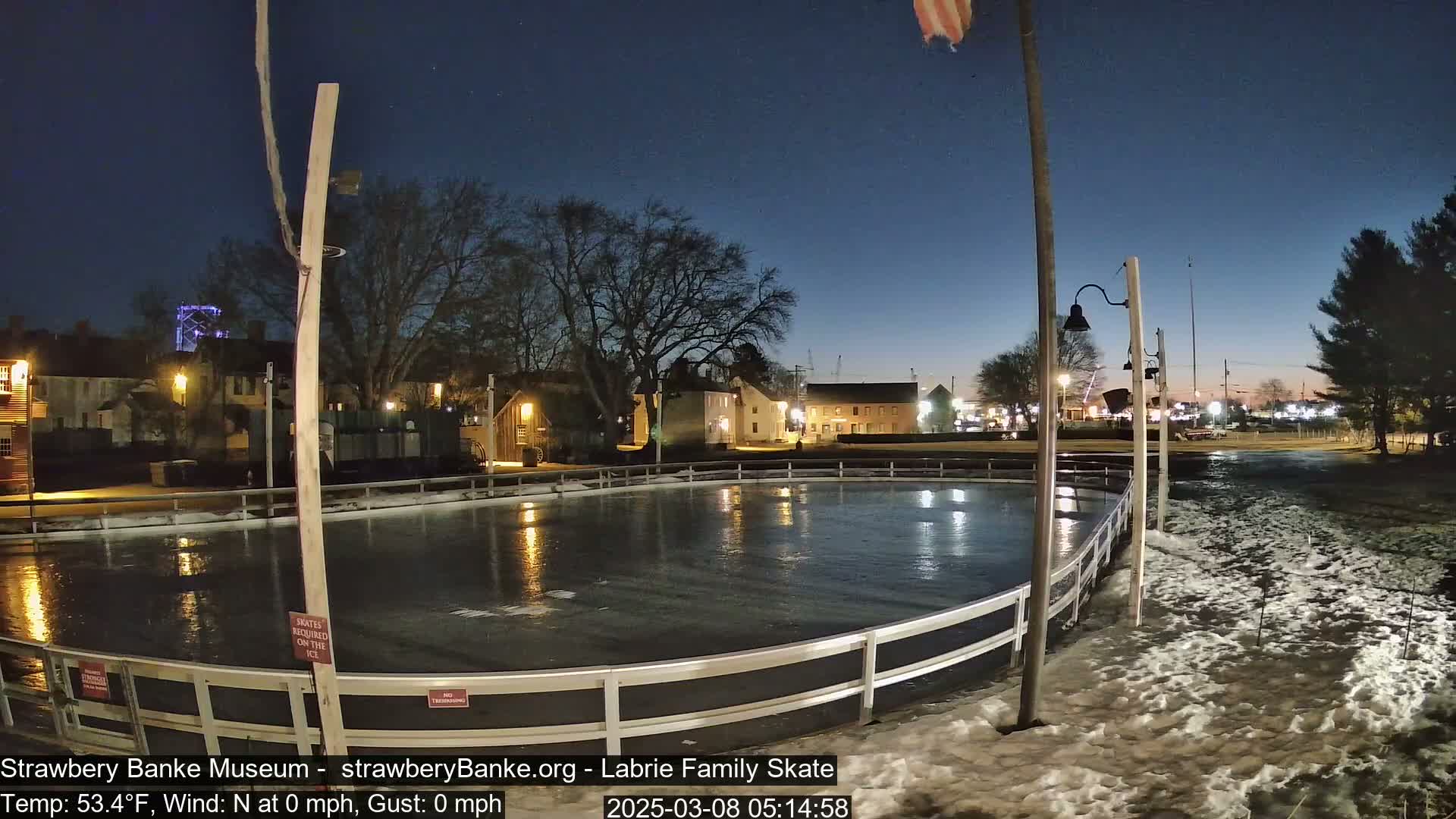 An outdoor ice skating rink at night under a twilight sky, with some snow on the ground near the rink and buildings visible in the background.