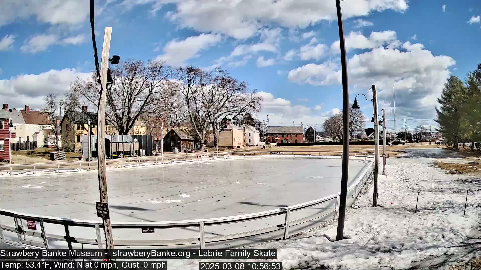 An outdoor ice skating rink is surrounded by snow and partially melted ground, with historical-looking buildings and trees in the background under a partly cloudy sky.