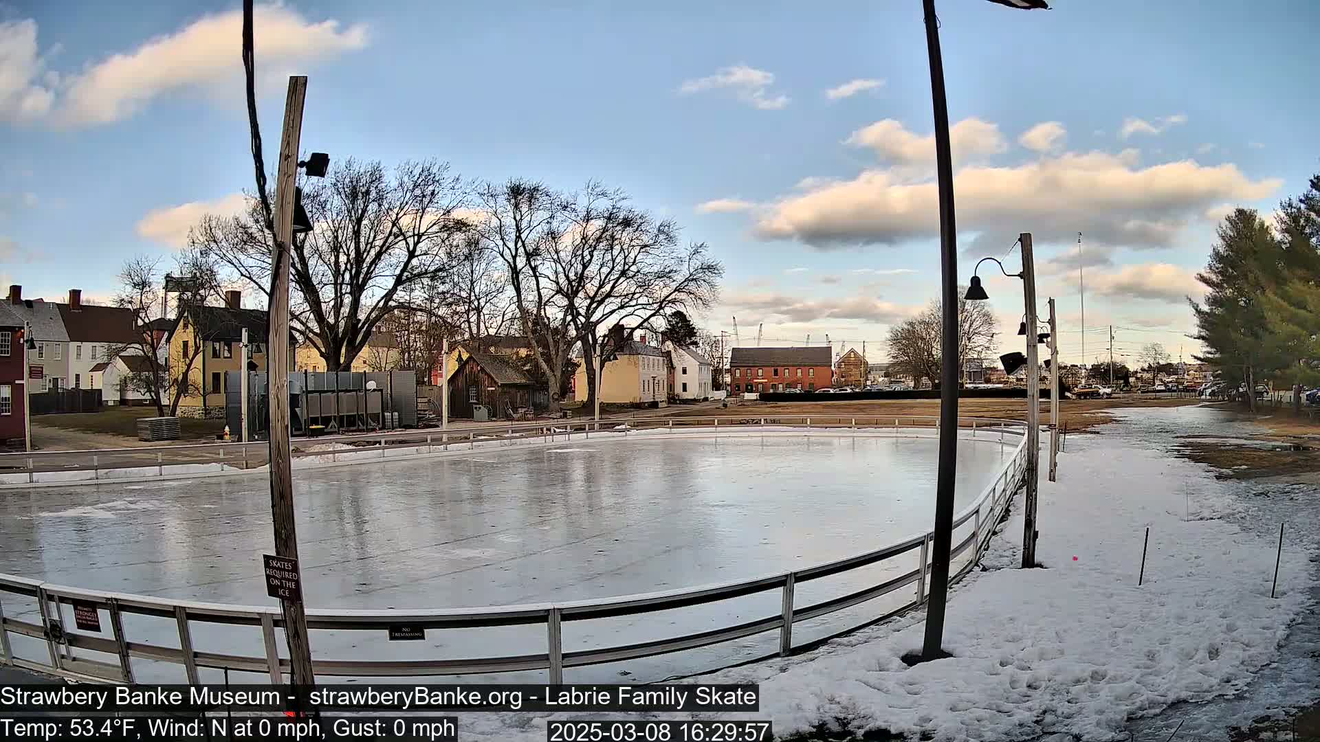 An outdoor ice rink sits beside several historic buildings under a partly cloudy sky.