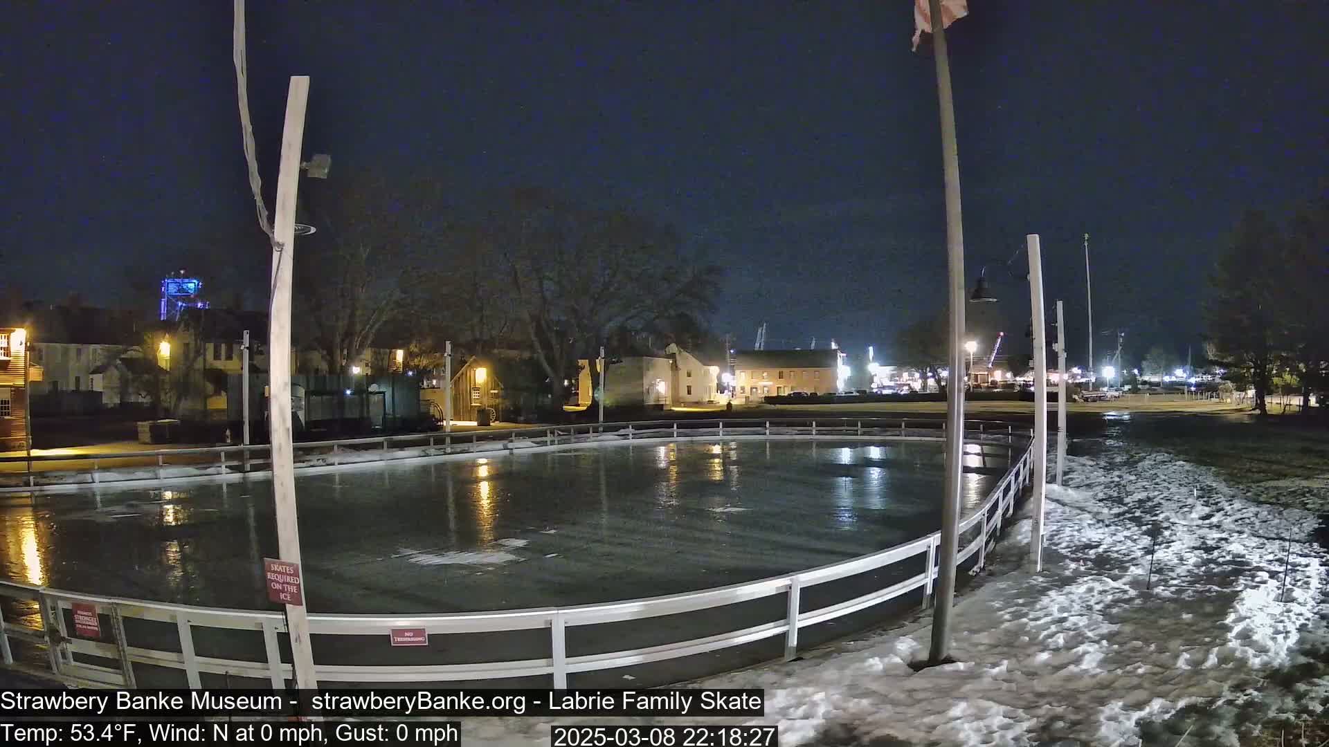 A nighttime view of an outdoor ice rink surrounded by snow, with calm weather conditions and buildings illuminated in the background.
