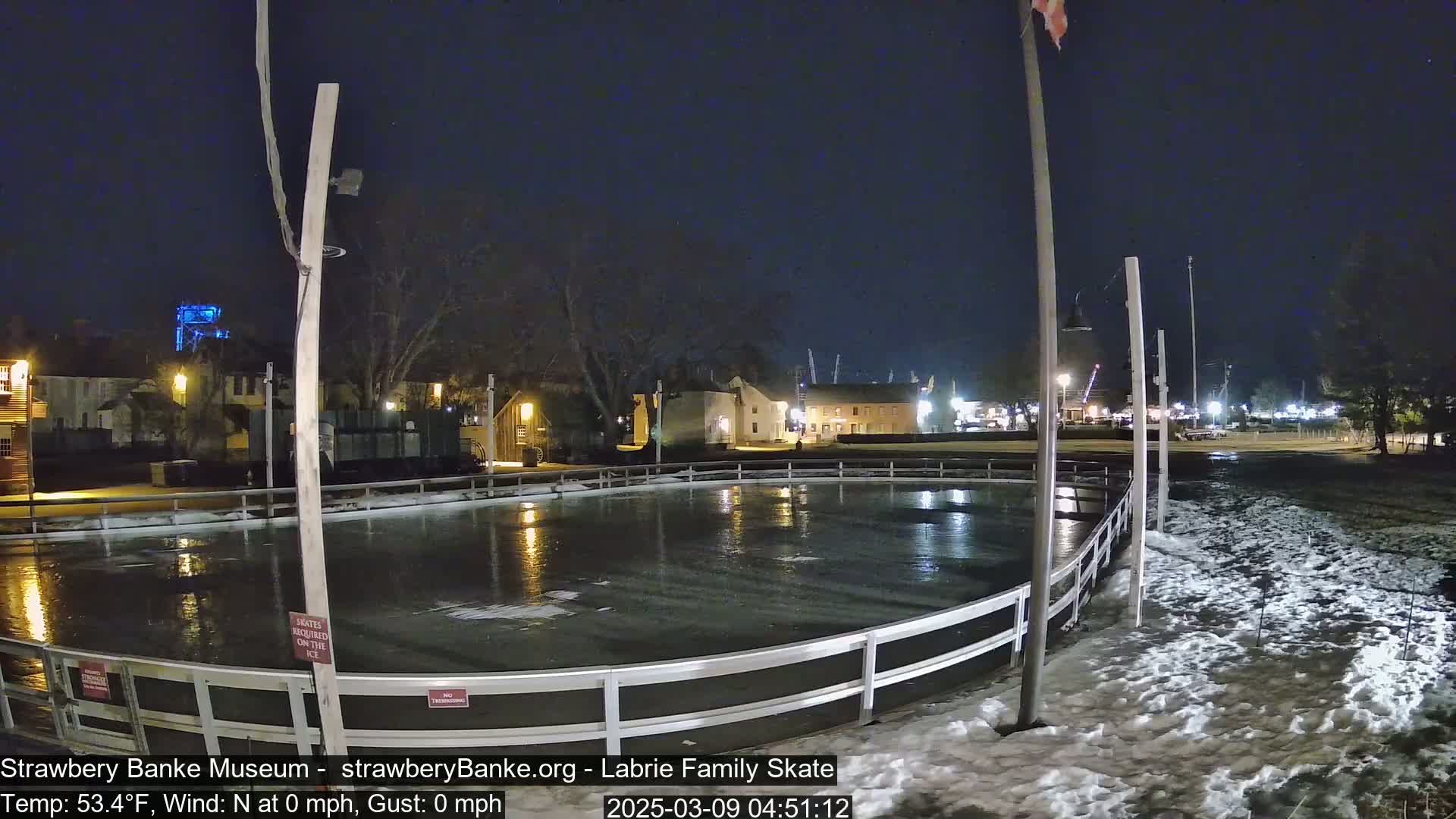 A nighttime view of an outdoor ice rink surrounded by snow, with buildings and streetlights visible in the background under a clear, dark sky.