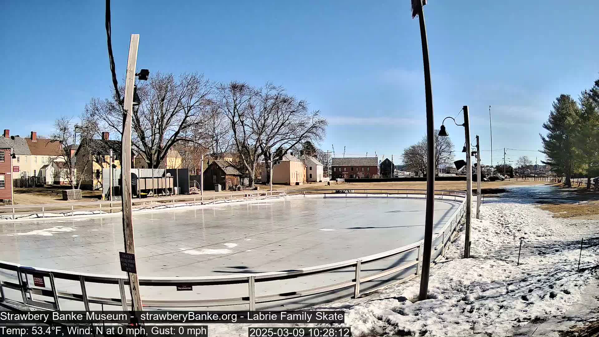 An outdoor ice skating rink, mostly clear of snow, is situated near several historic-looking buildings under a clear blue sky.