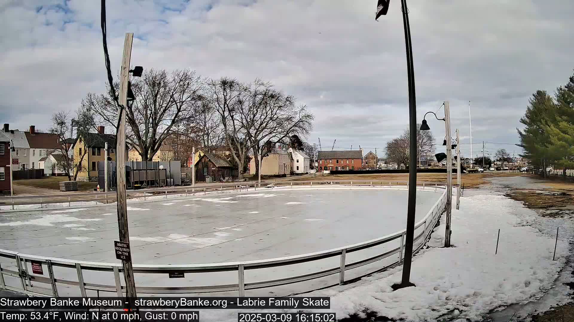 An outdoor ice rink is surrounded by patchy snow, some buildings, and bare trees under a cloudy sky.