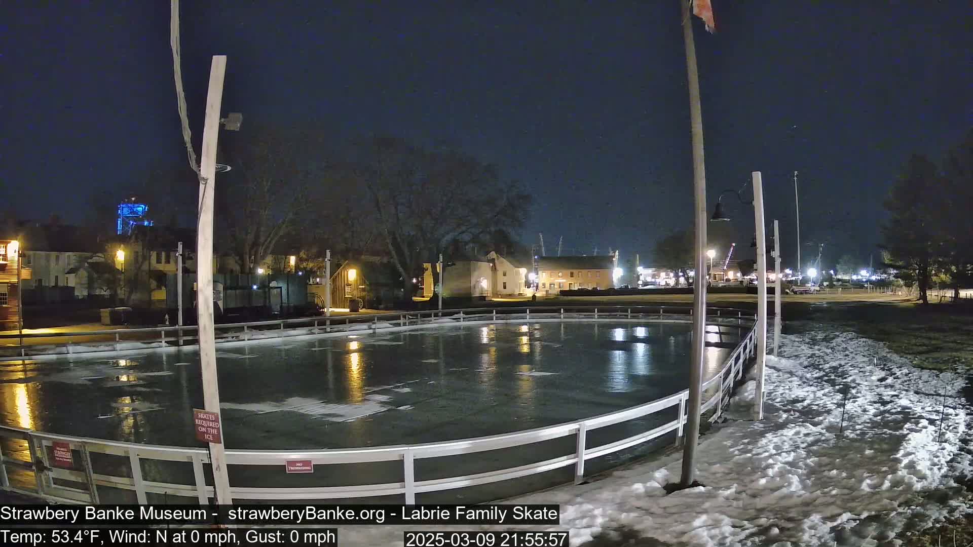 An outdoor ice rink at night is illuminated, surrounded by snow, with calm conditions and nearby buildings faintly visible under a dark sky.