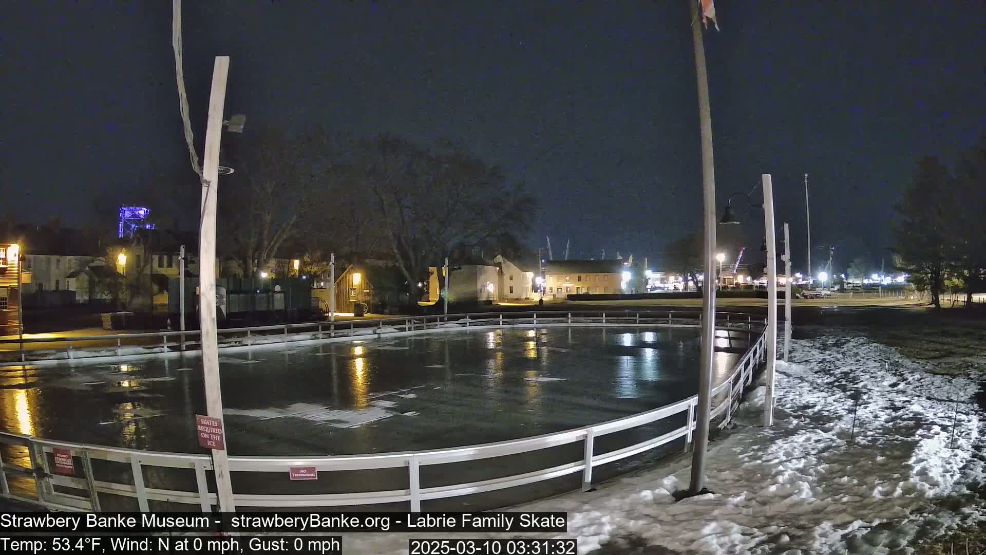 A nighttime view of an outdoor ice rink, partially surrounded by snow, with calm conditions and buildings in the background.