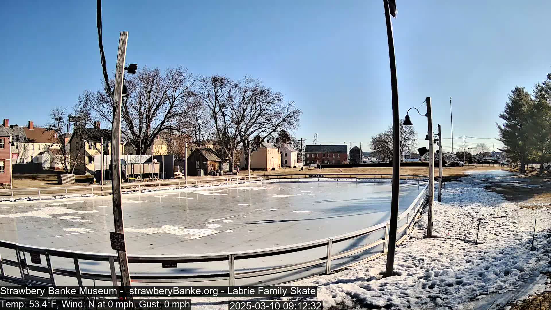 An outdoor ice rink, surrounded by snow and bare trees, sits in front of historical-looking buildings under a clear blue sky.
