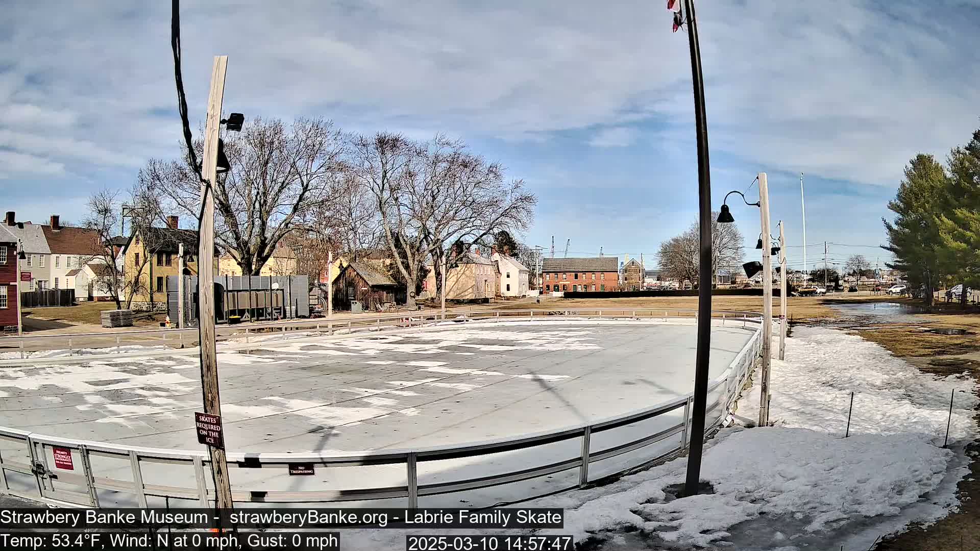 An outdoor ice rink sits partially surrounded by snow and partially melted ground, under a mostly sunny sky, near several historical-looking buildings.