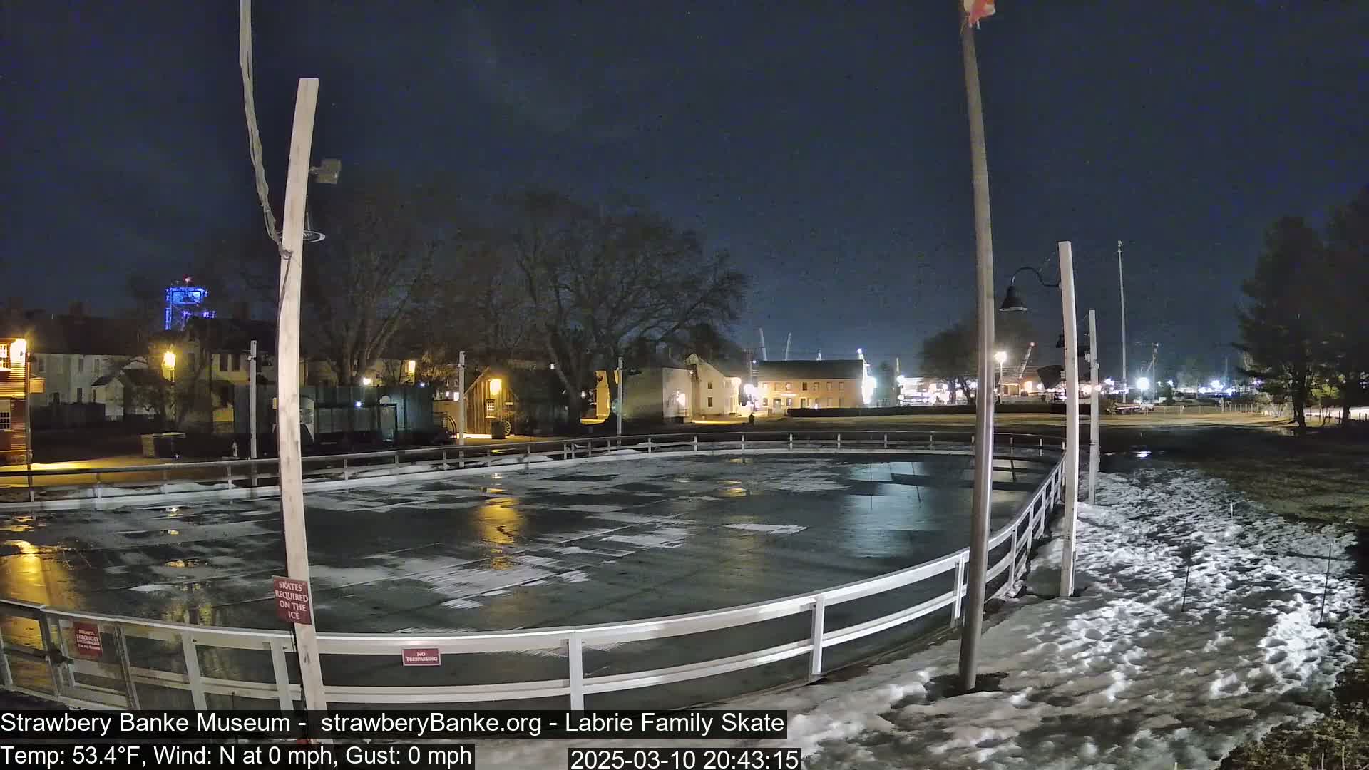 An outdoor ice rink at night, surrounded by snow and lit buildings, under a clear, dark sky.