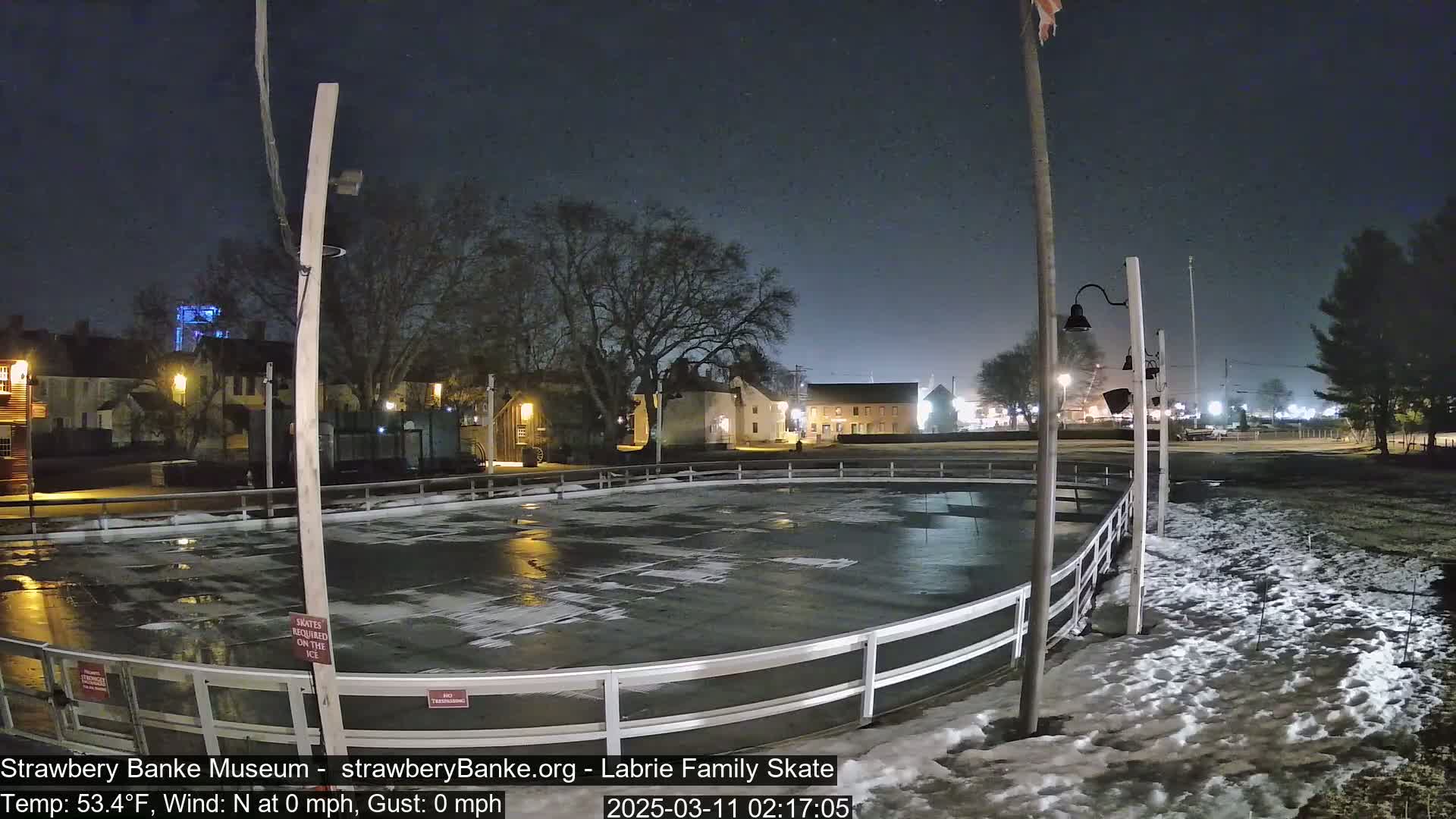 An outdoor ice rink, surrounded by a low white fence, is illuminated at night under a clear, dark sky with some patchy snow on the ground.