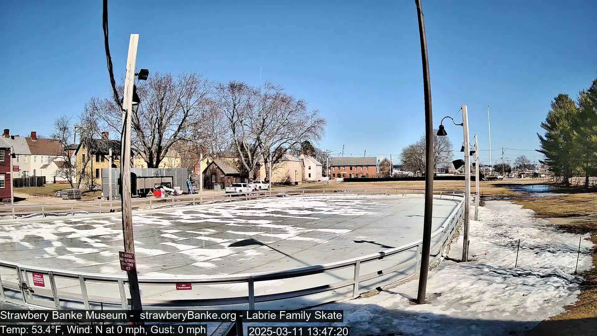 An outdoor ice skating rink, partially surrounded by melting snow, sits in front of a cluster of historic-looking buildings under a clear blue sky.