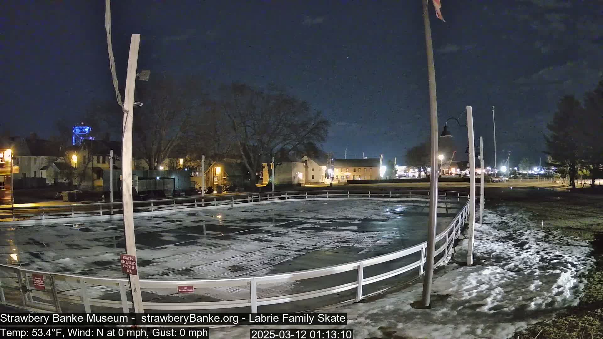 An outdoor ice rink sits in front of a row of buildings at night, with a calm, clear sky and a small amount of snow on the ground.