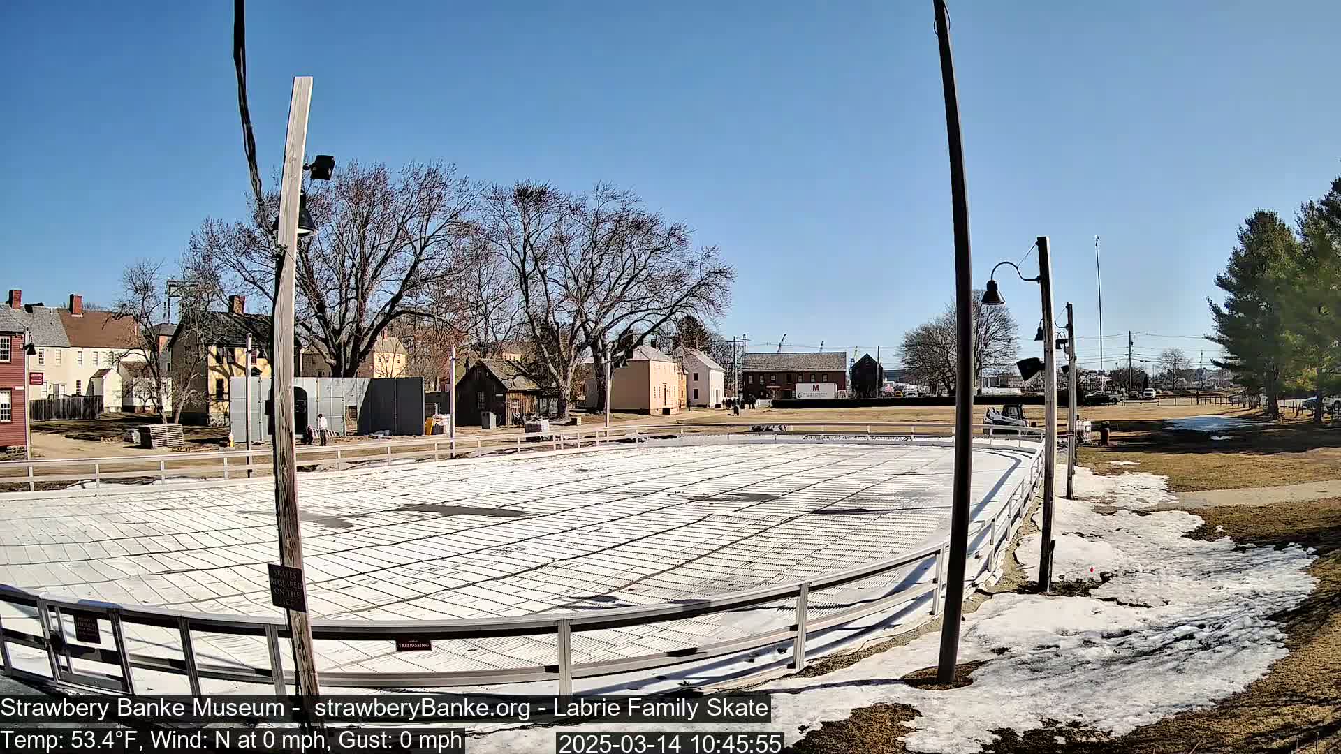 A mostly clear day shows an empty outdoor skating rink surrounded by historical-looking buildings and patchy snow.