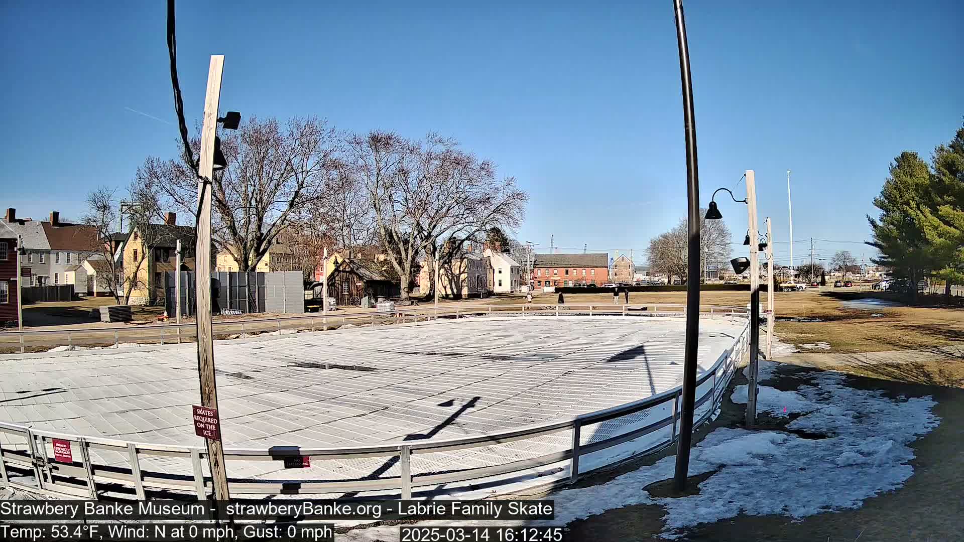 An outdoor ice skating rink, surrounded by patchy snow and brown grass, sits in front of several buildings under a clear blue sky.