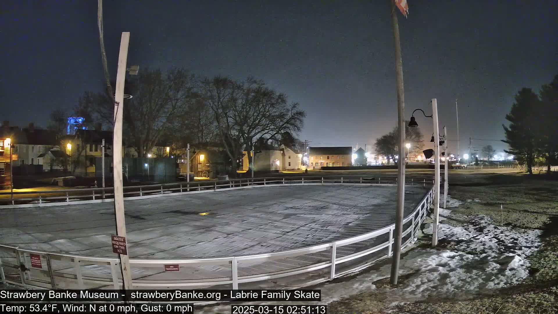 A nighttime view of an outdoor ice rink, partially covered in snow, surrounded by buildings and trees under a clear, dark sky.