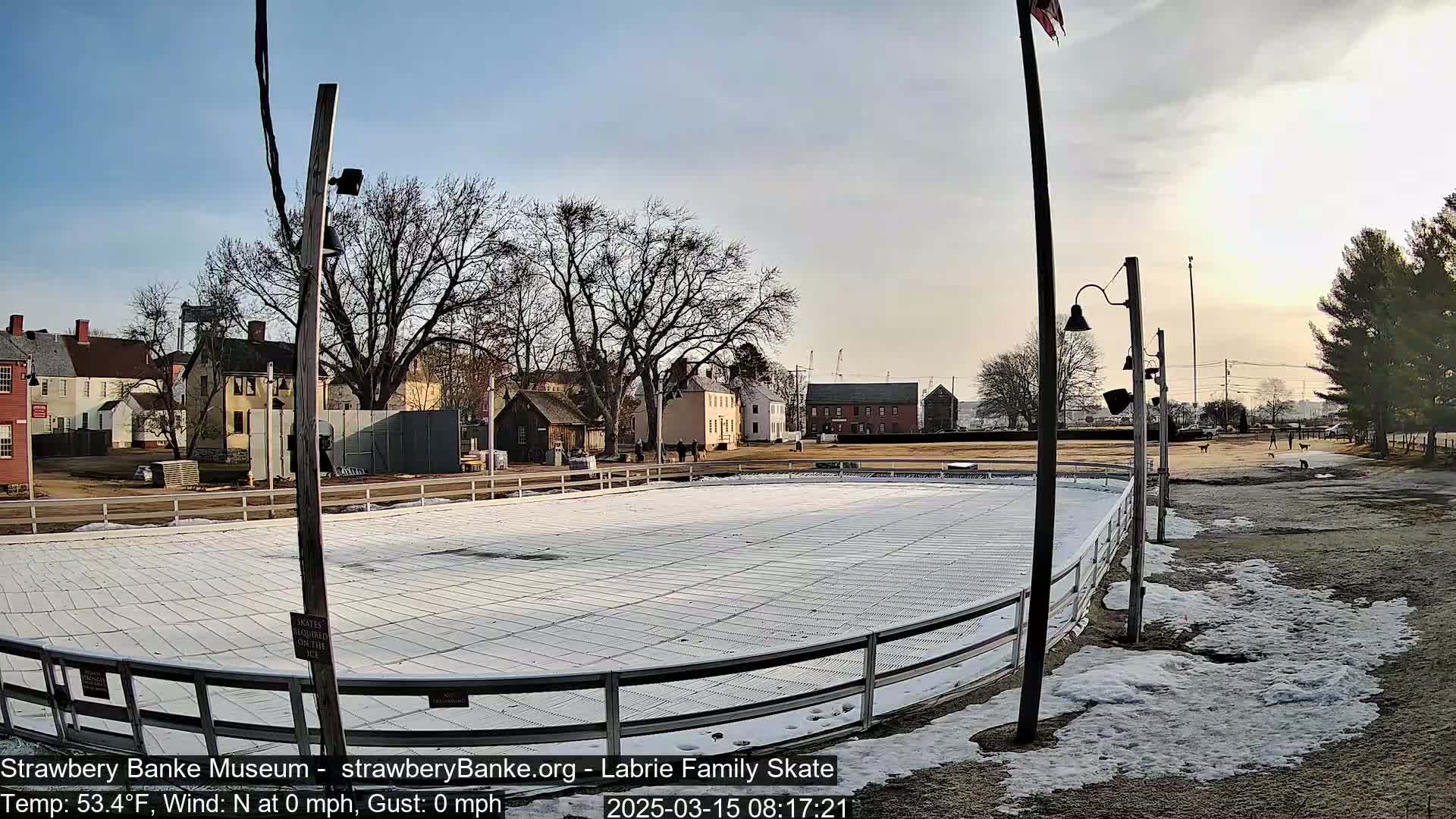 An outdoor ice skating rink, mostly covered in white ice, sits near several historical-looking buildings under a mostly clear sky with a slight amount of snow on the ground around the rink.