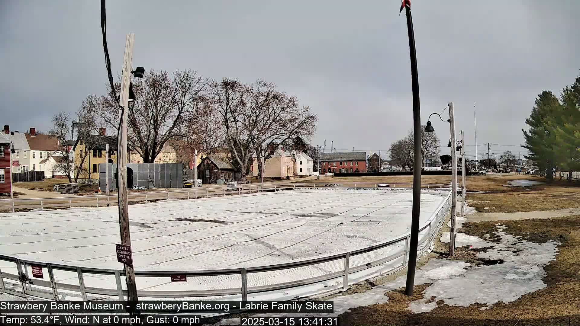 An outdoor ice rink sits empty in front of several colonial-style buildings on a partly cloudy day with no wind.