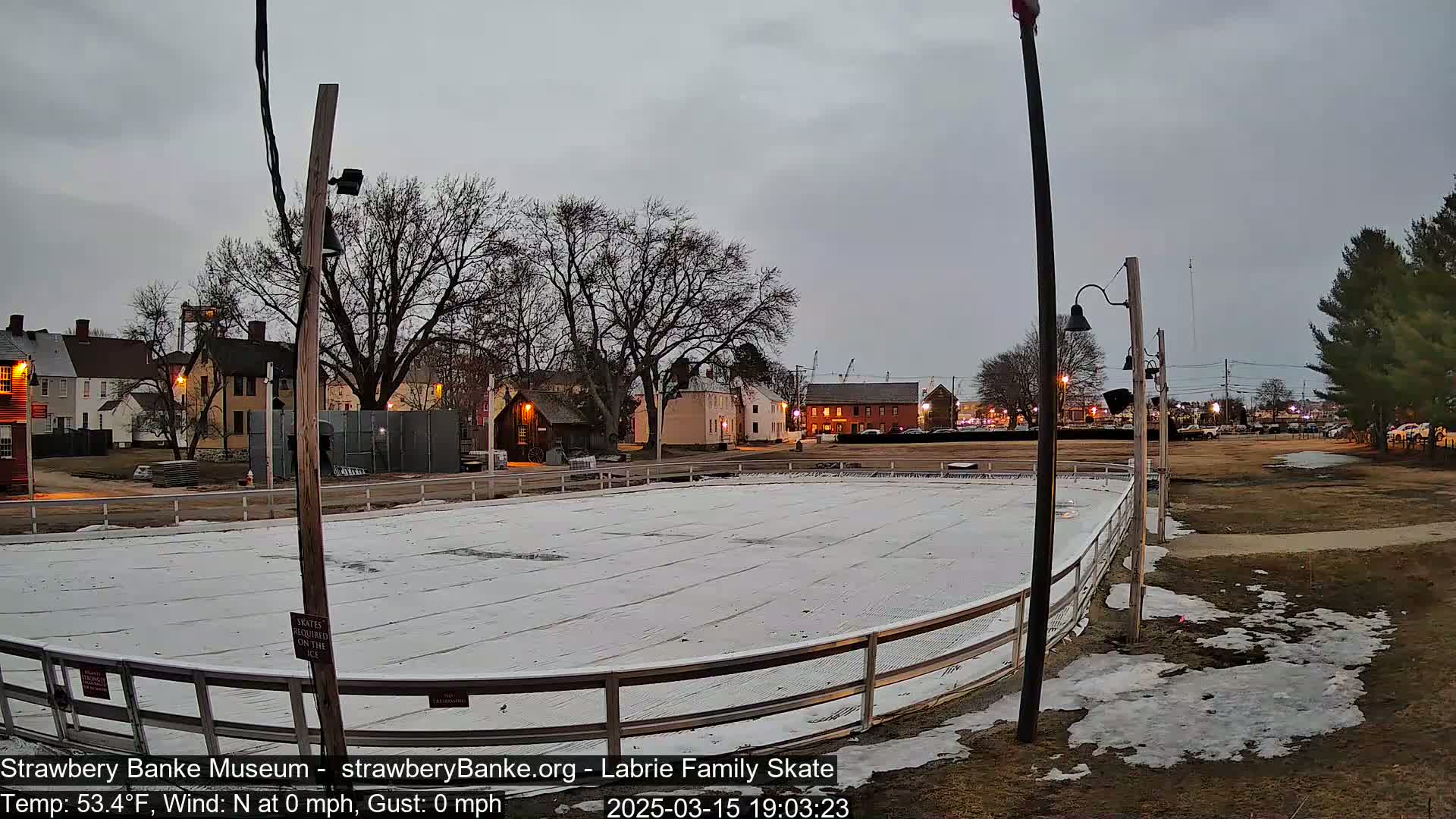 A snow-covered outdoor skating rink is situated in front of several historic-looking buildings under a cloudy, overcast sky.