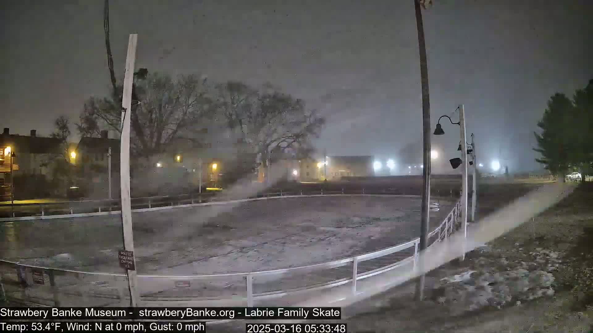 A snow-covered outdoor skating rink is mostly empty under a dark, stormy sky at night.