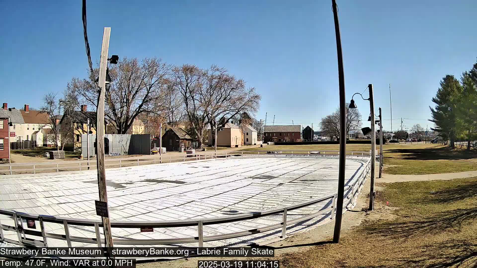 An outdoor skating rink, seemingly empty, sits beside several historic-looking buildings under a clear, sunny sky.