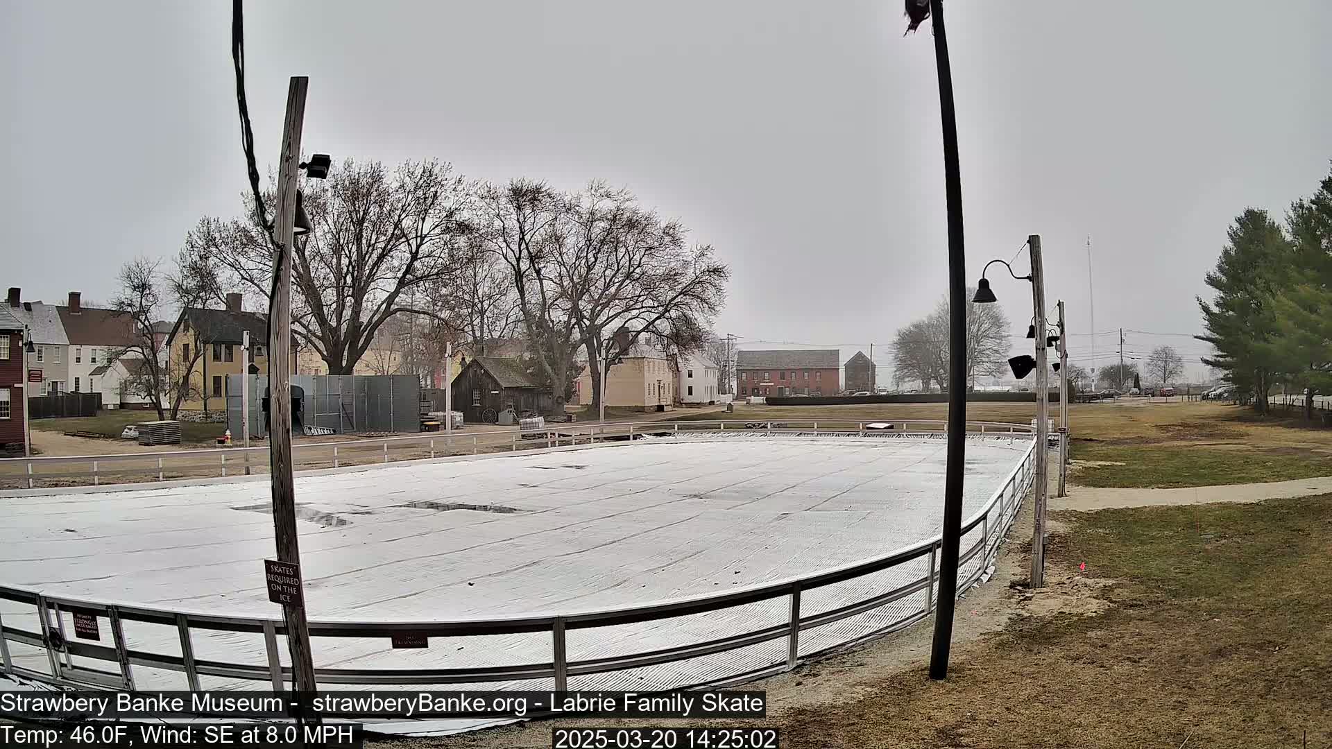 A covered outdoor ice skating rink sits in front of several historic-looking buildings on an overcast day with a southeast wind.