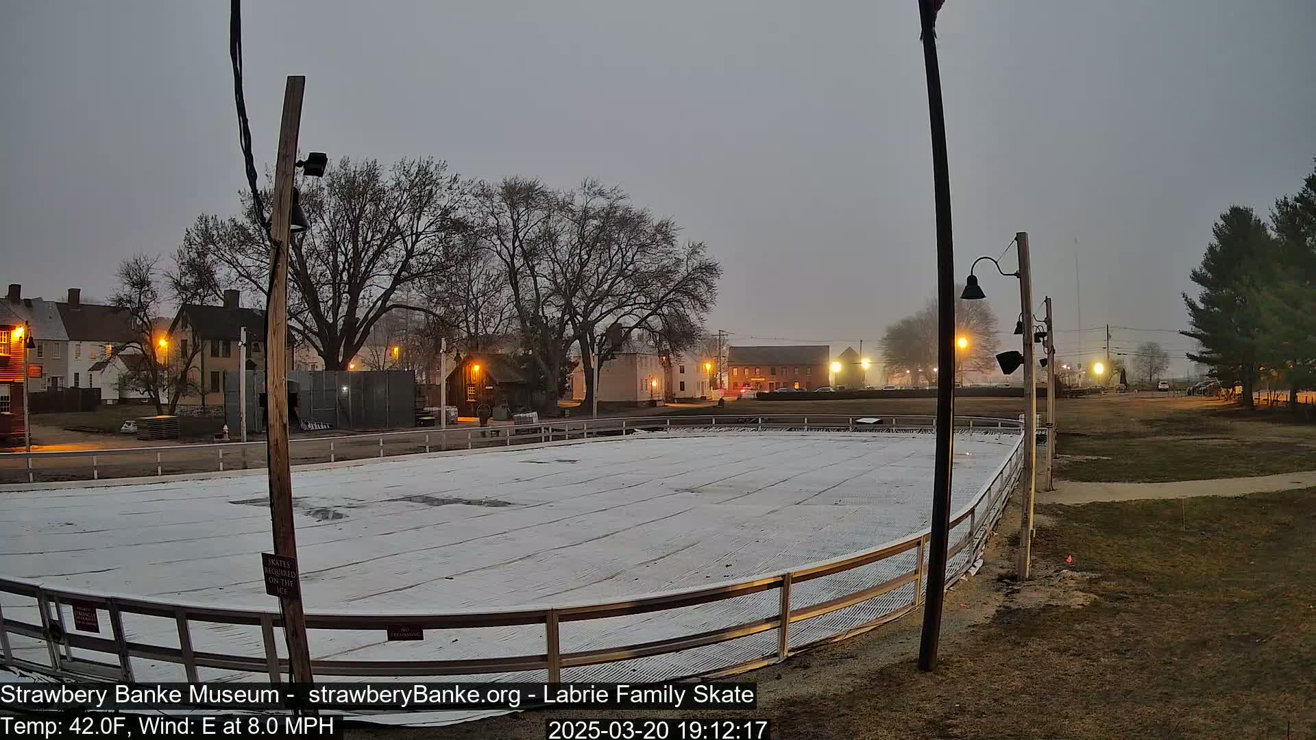 A covered outdoor ice rink sits in a dimly lit town square under a foggy, twilight sky.