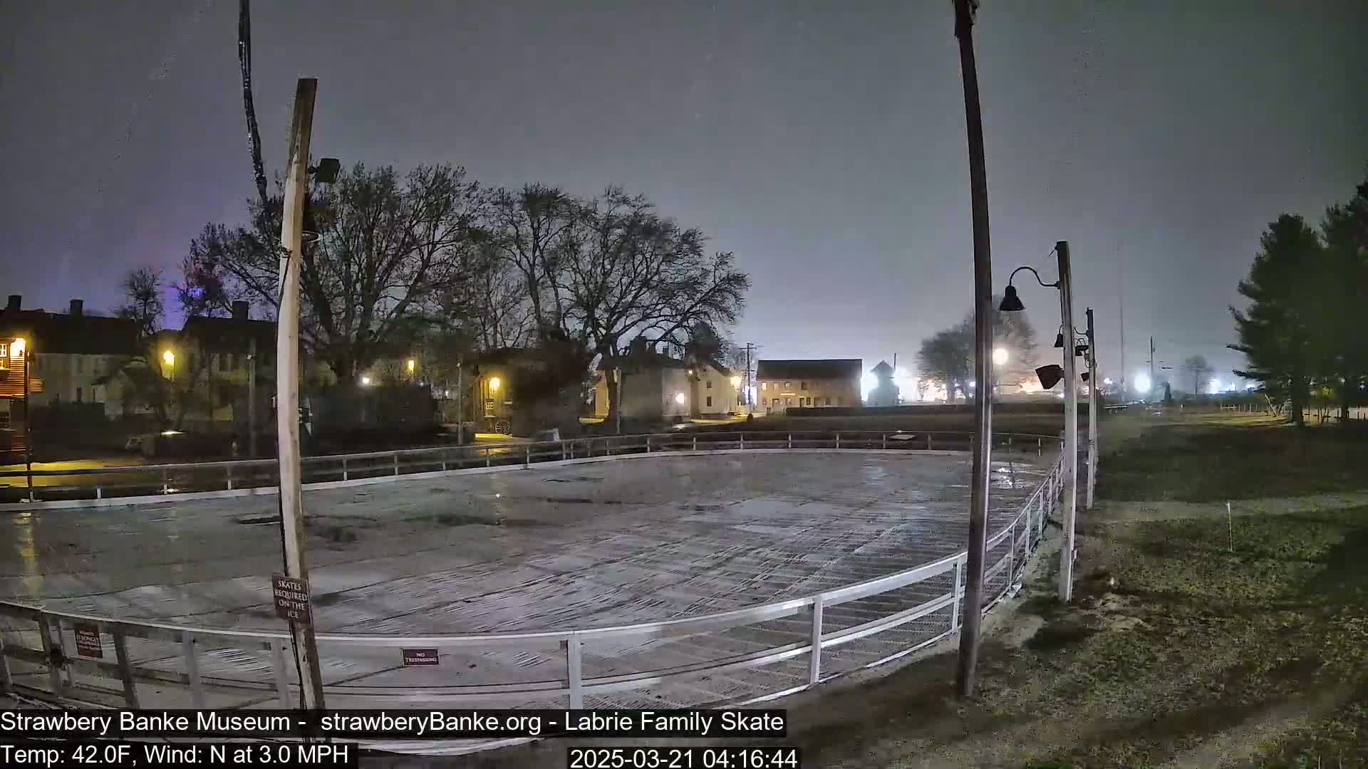 An empty outdoor ice rink is visible at night under a hazy sky with a light north wind.