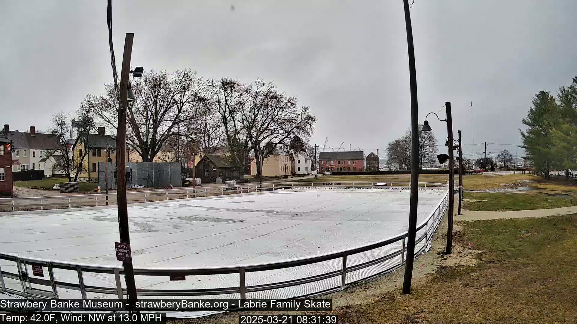An outdoor ice rink sits beside a row of historic buildings on an overcast day with a northwest wind.
