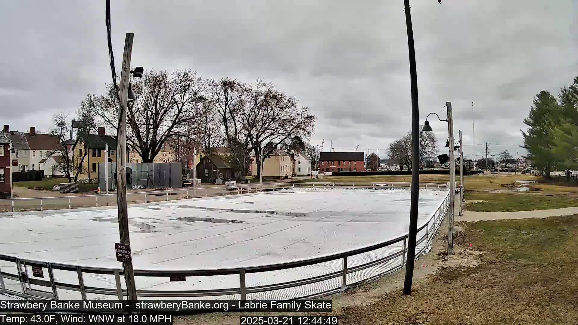 An outdoor ice rink sits in front of historical-looking buildings under an overcast sky, with a wind of 18 mph.